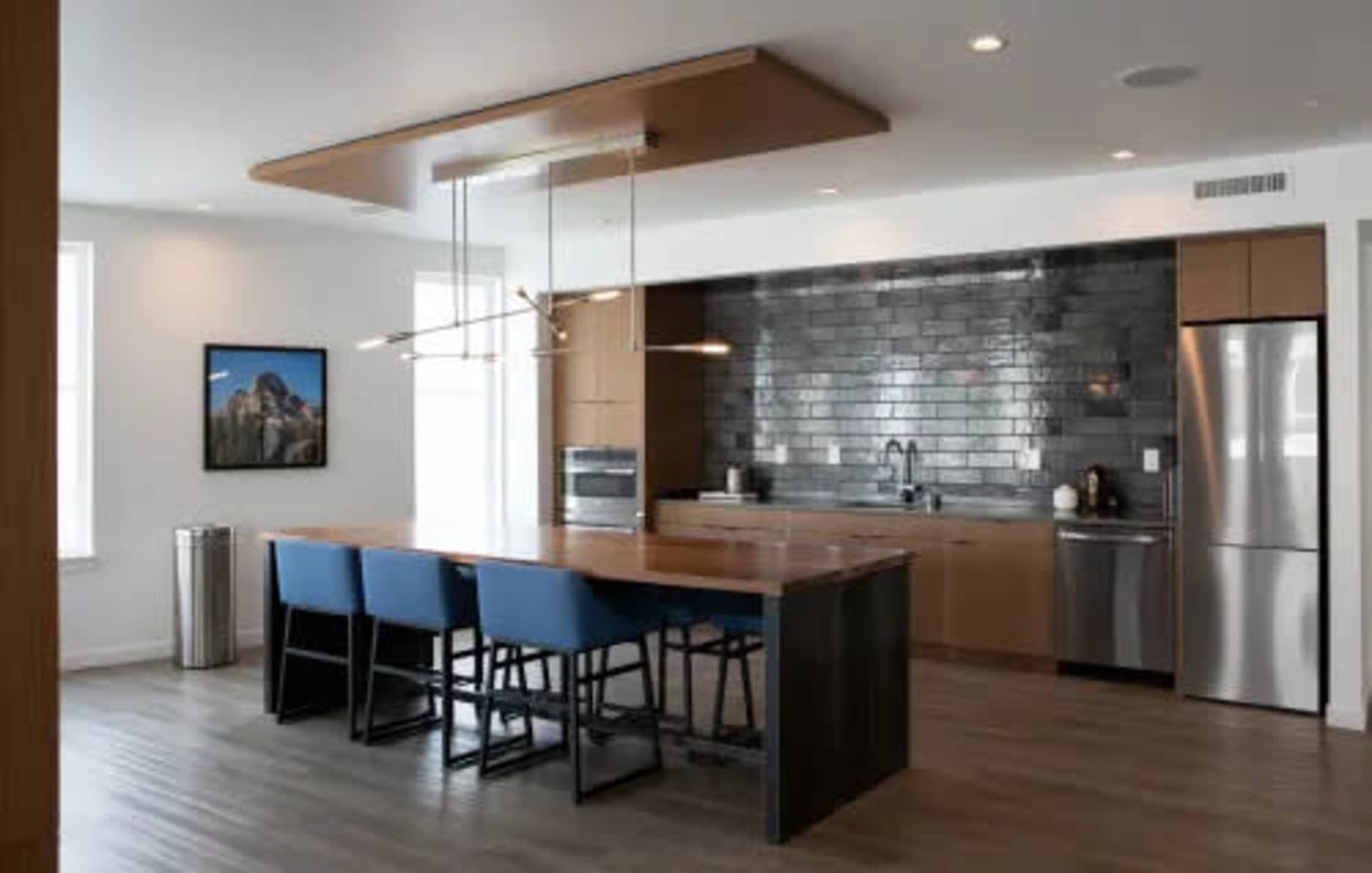 A modern kitchen features a large island with blue chairs, stainless steel appliances, and dark tiled backsplash.