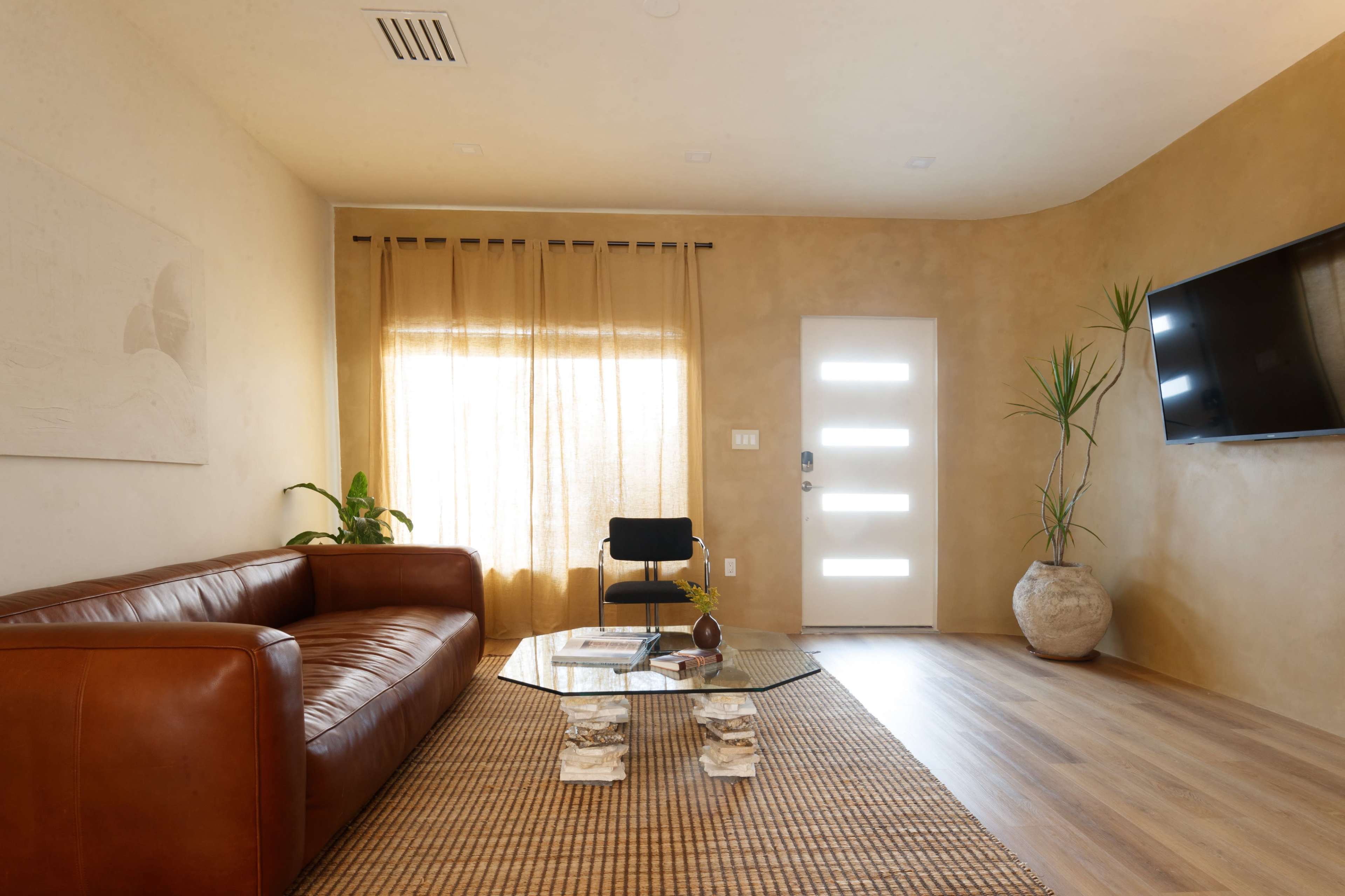 A modern living room features a brown leather sofa, a glass coffee table, large windows with sheer curtains, and a minimalist décor style.