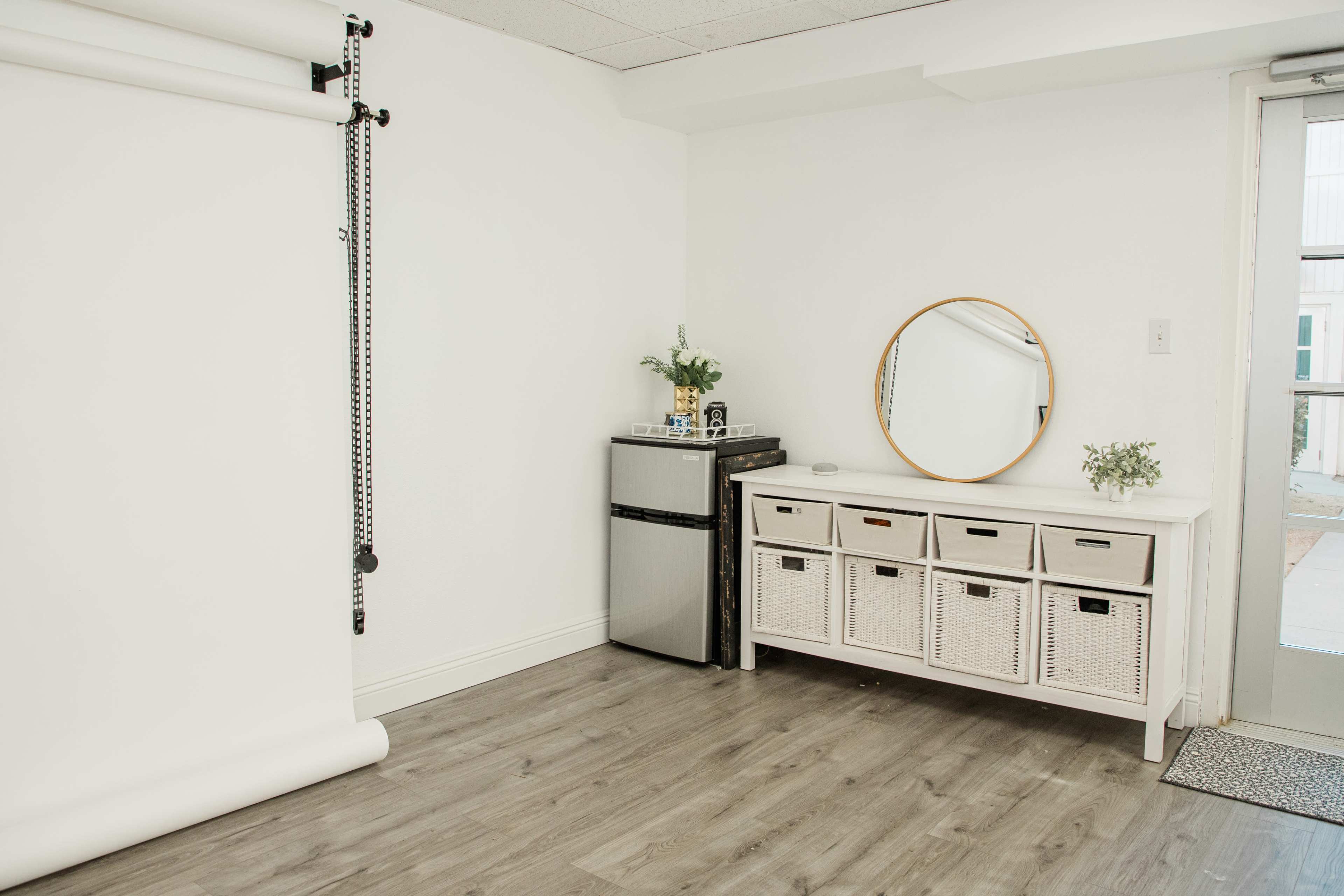 The image shows a minimalist room with a gray wooden floor, a white wall, a small refrigerator, a console table with woven baskets, and a round mirror.