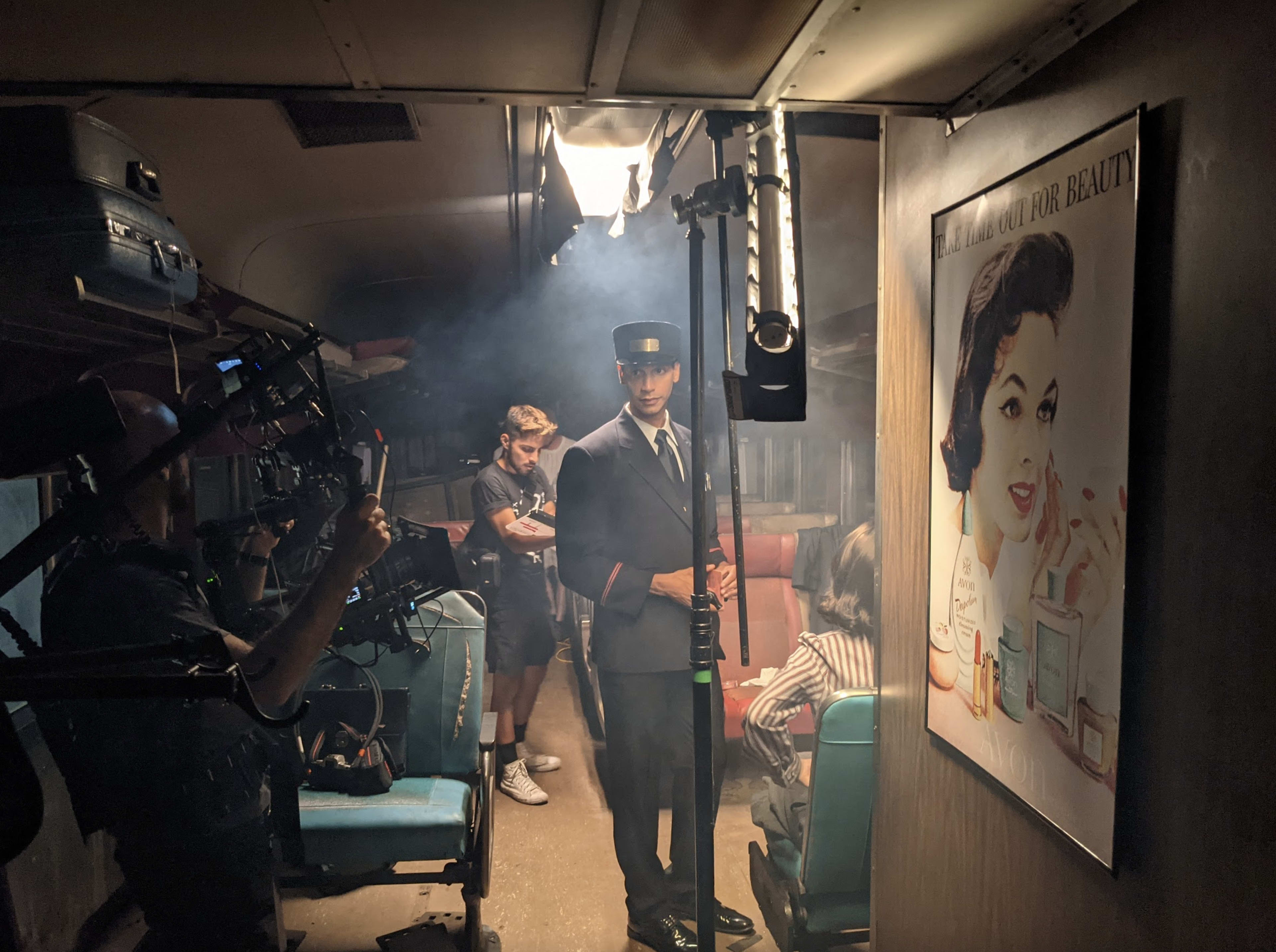 A film crew works on a set designed to resemble an old train car, featuring a conductor in uniform and an advertisement on the wall.