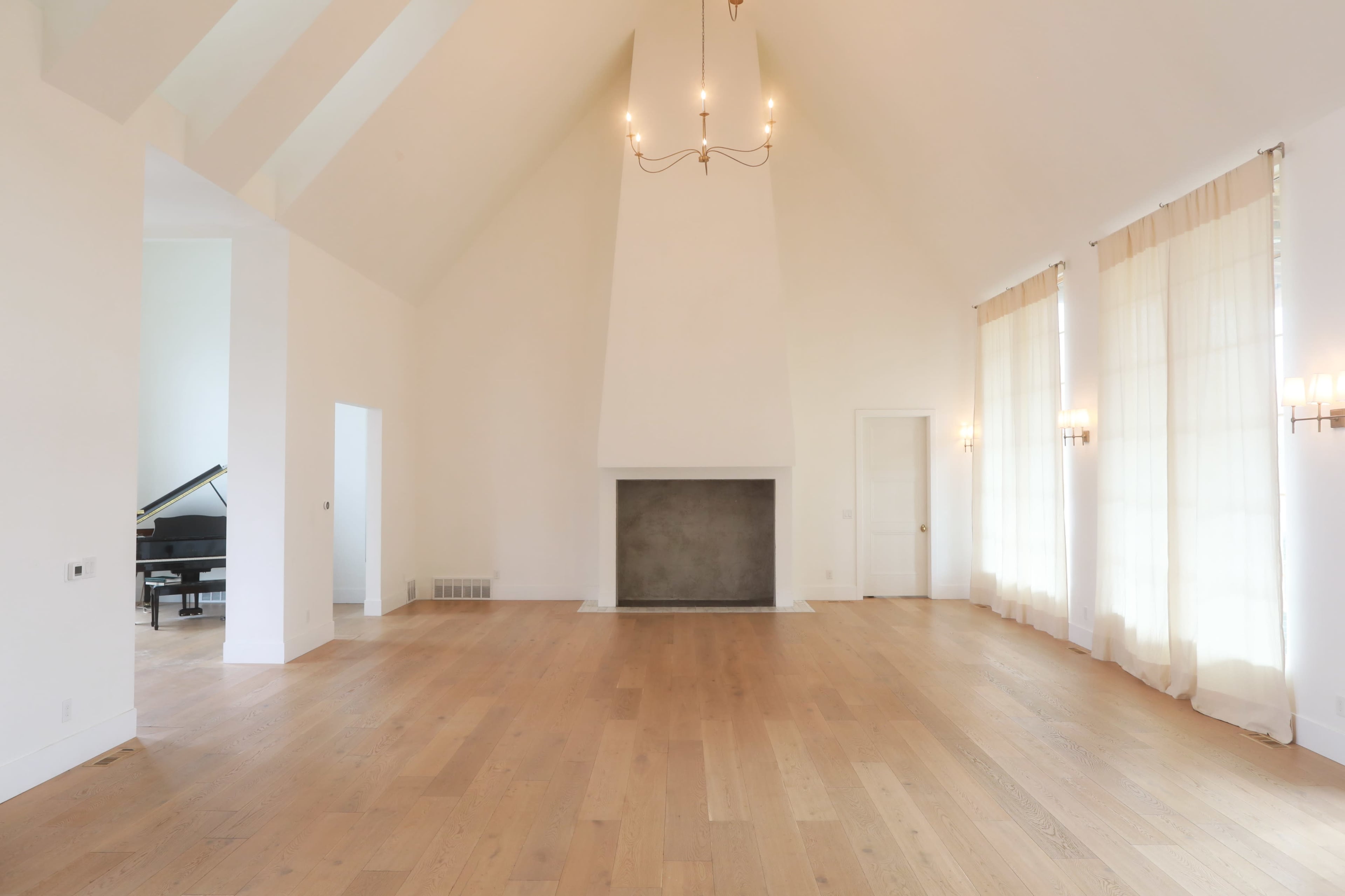 The image shows a spacious interior living area with high ceilings, a fireplace, large windows covered by sheer curtains, and a grand piano in the background.