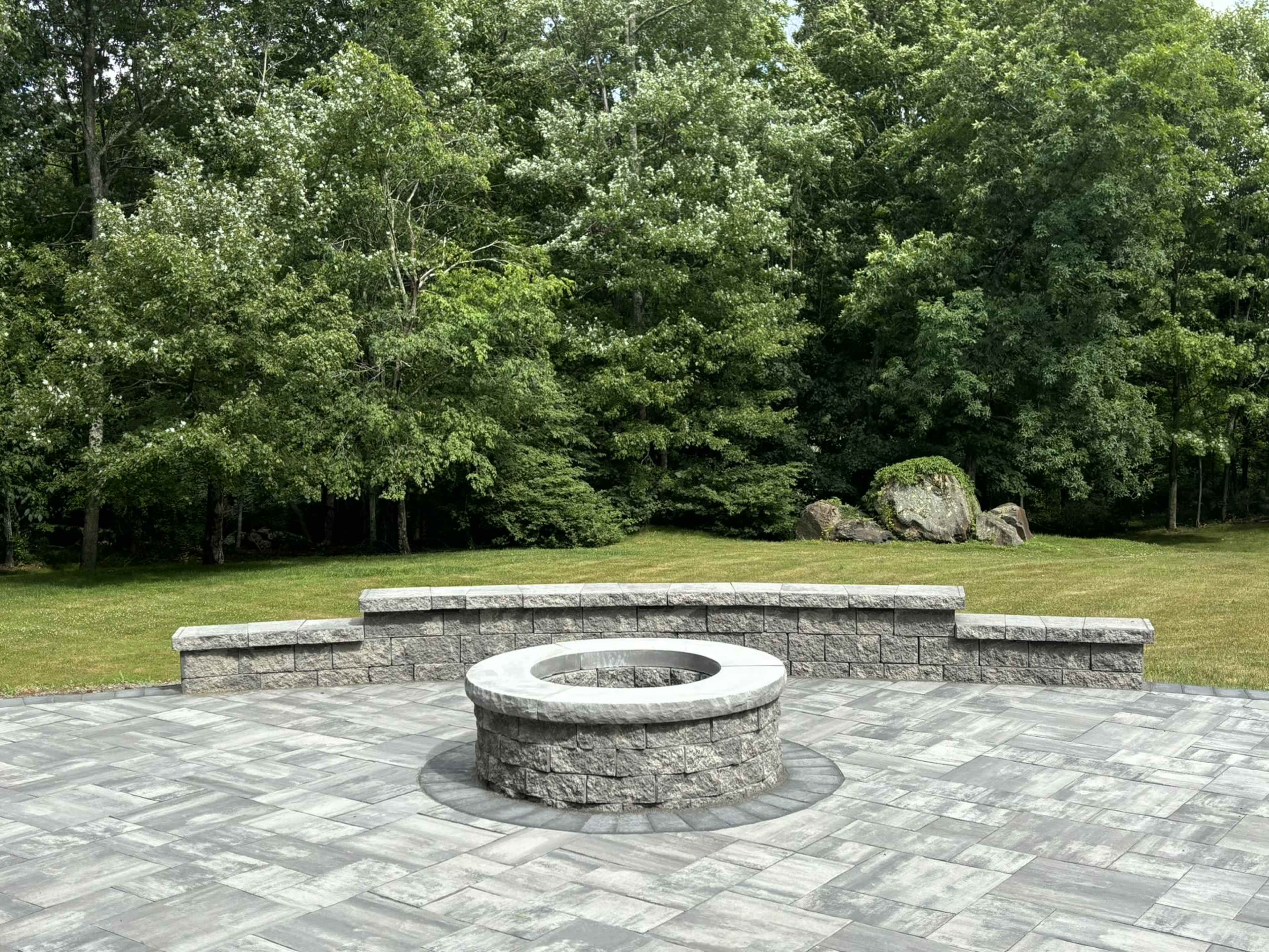 A circular stone fire pit is set in the center of a stone patio surrounded by lush green trees.