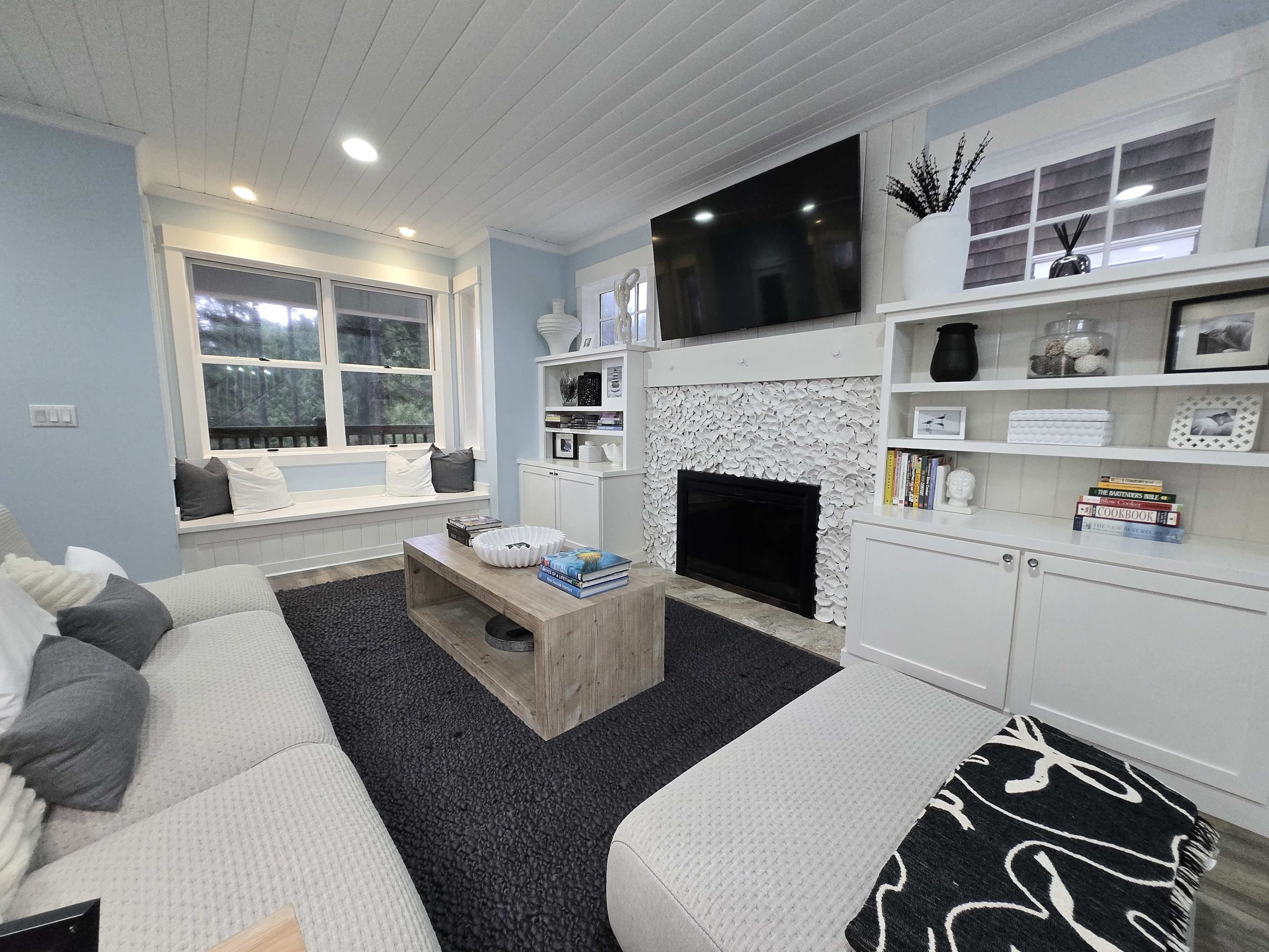 The image shows a cozy living room featuring a light-colored sofa, a wood coffee table, a fireplace with a stone facade, built-in shelves, and a large flat-screen TV mounted on the wall.