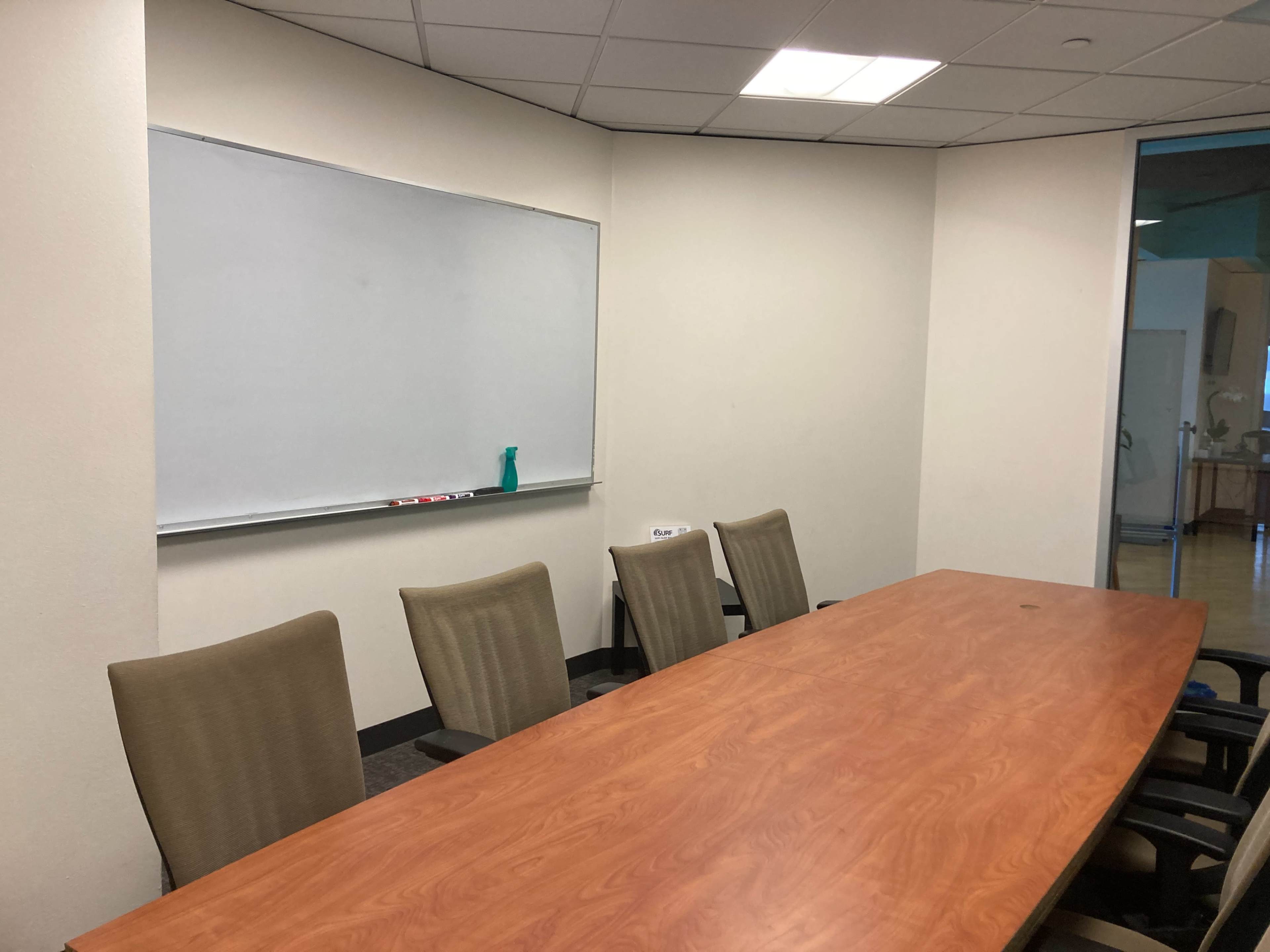 A conference room features a long wooden table surrounded by five chairs and a whiteboard on the wall.