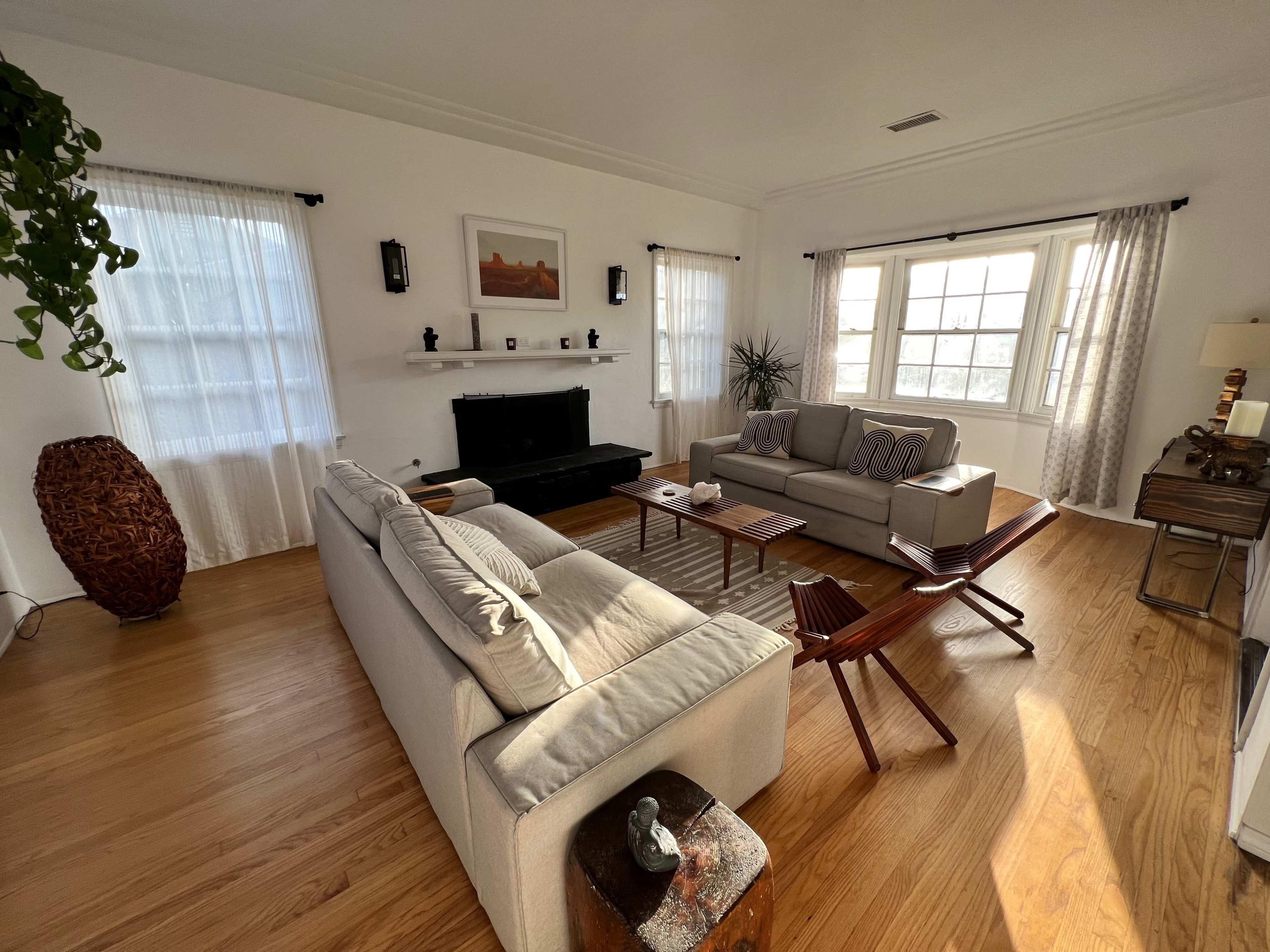 A bright living room features a light-colored sofa, two armchairs, a coffee table, and a fireplace, all set against large windows and wooden floors.