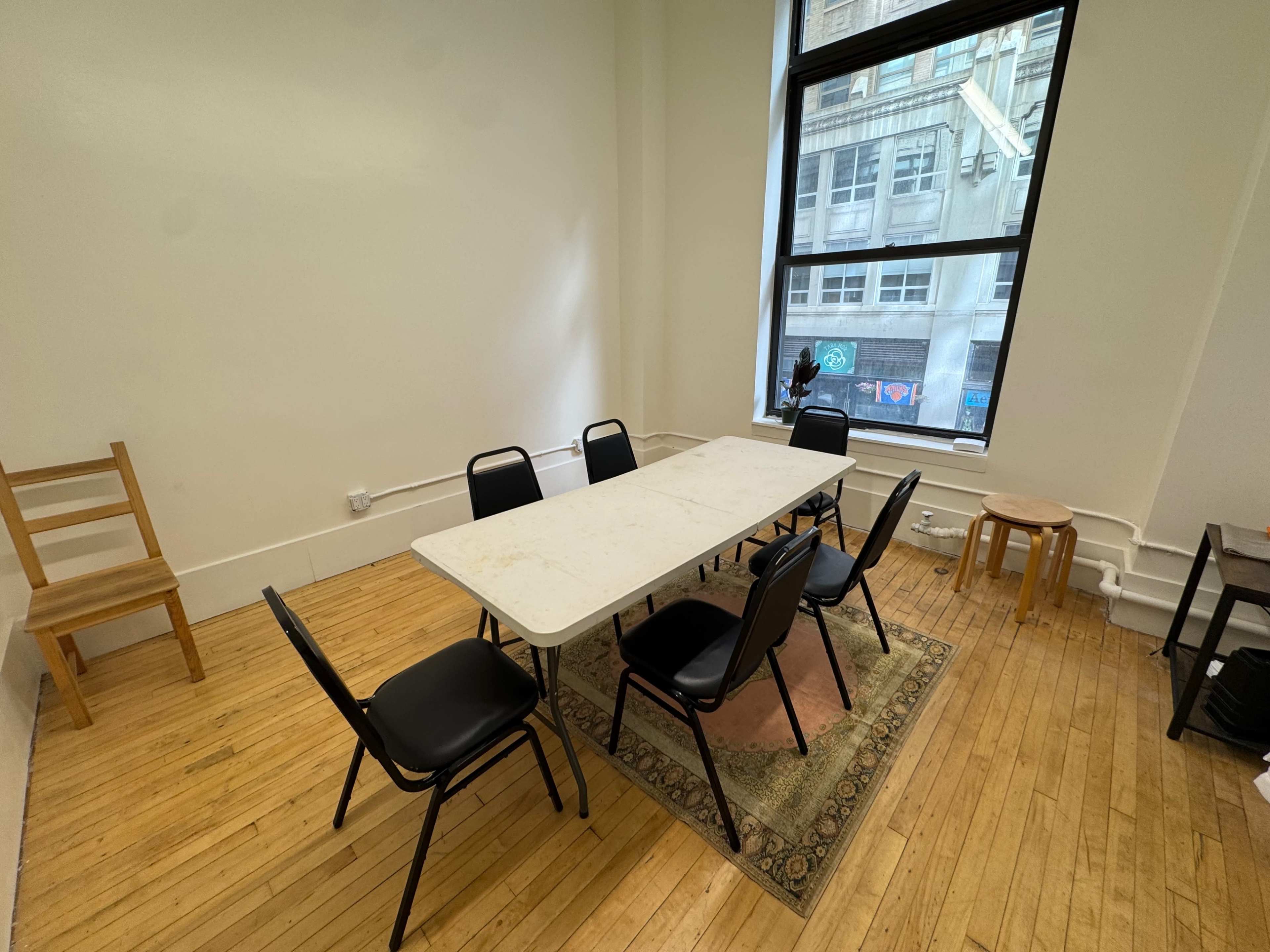 The image shows a minimalist room with a rectangular table surrounded by six chairs, a wooden chair in the corner, and a small stool near a window.