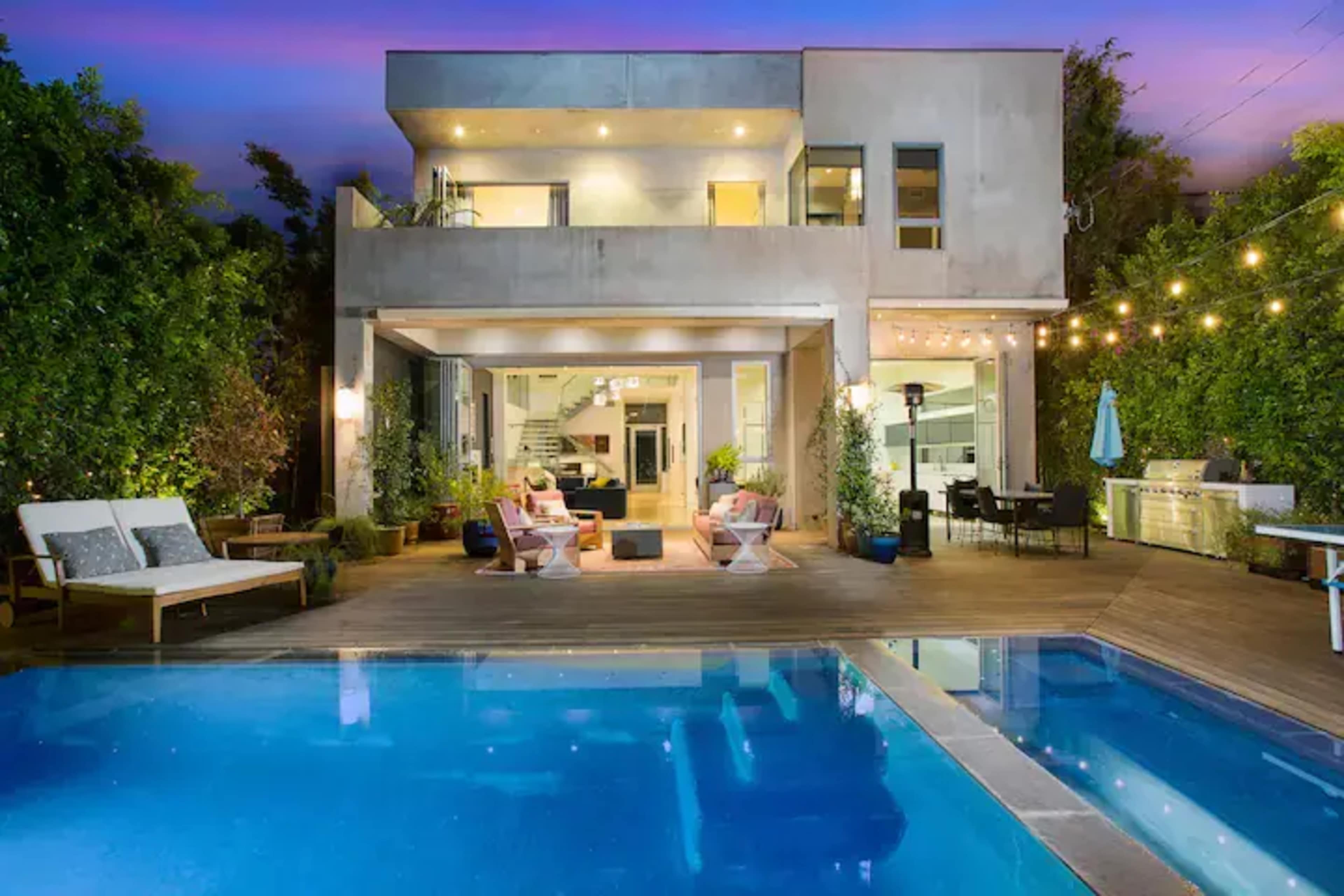 A modern two-story house with a swimming pool and outdoor seating area, illuminated by string lights at dusk.