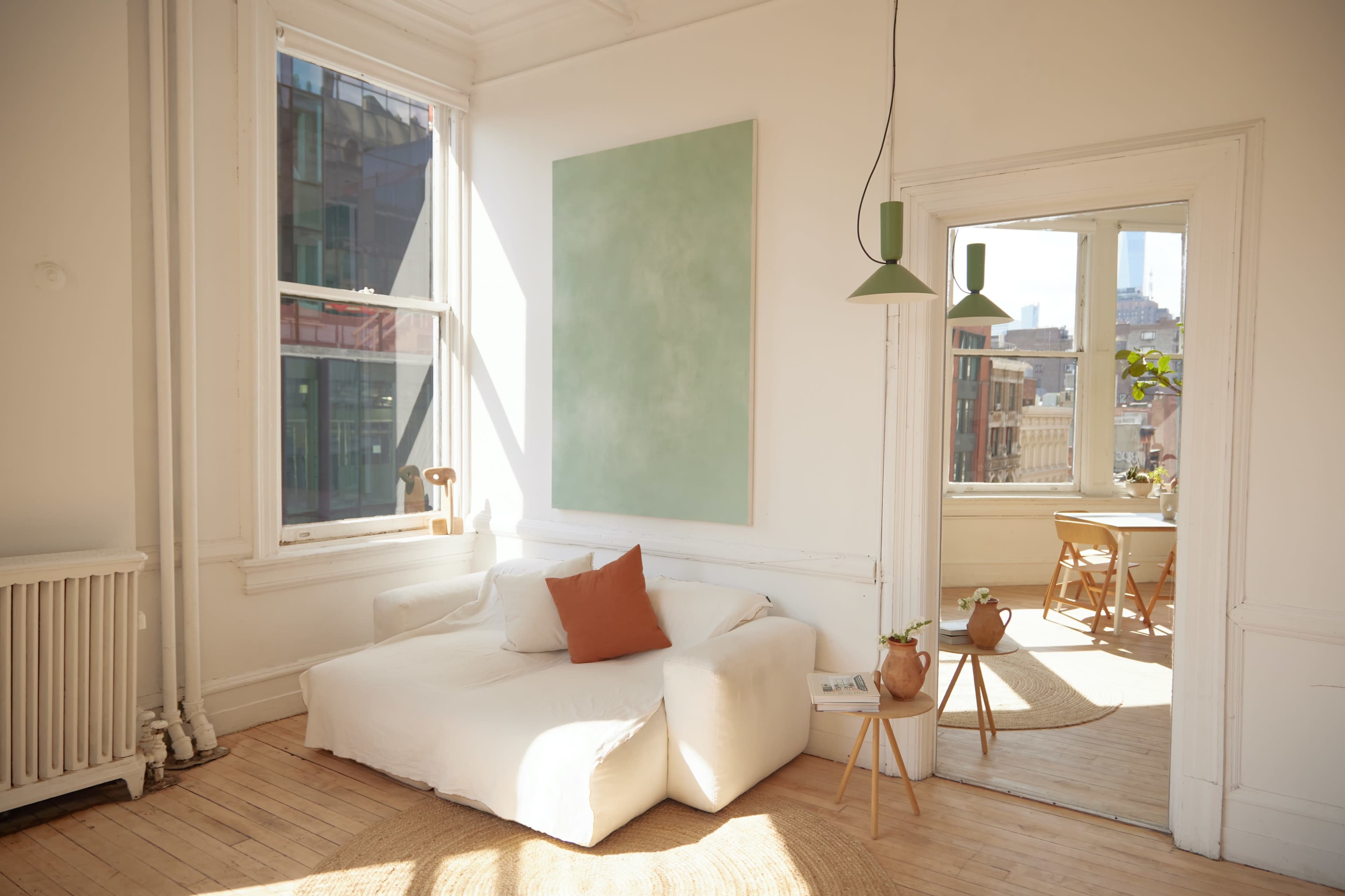 A bright, minimalistic living room features a white sofa with an orange cushion, large windows, and an adjacent dining area visible through an open door.