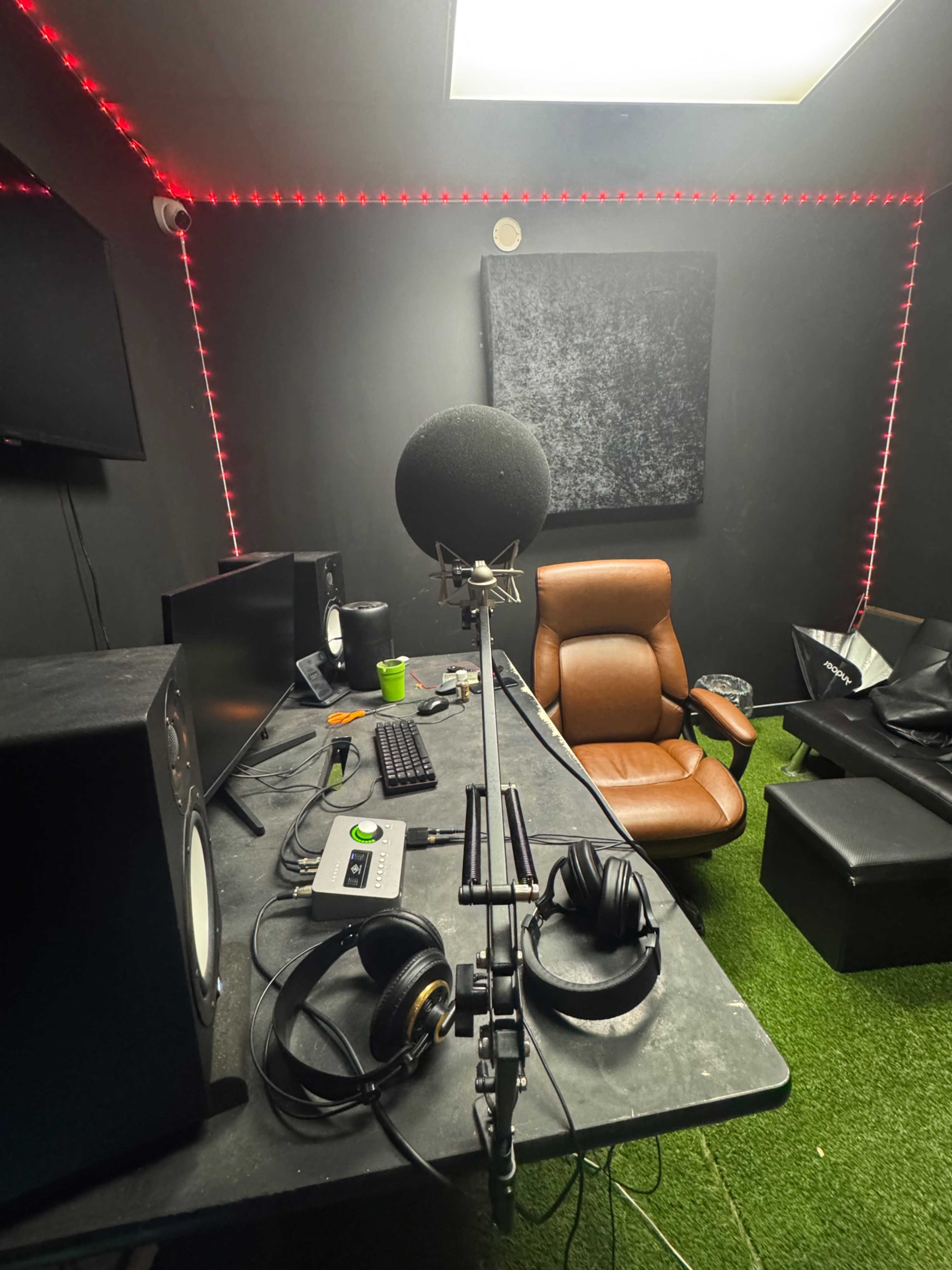 The image shows a recording studio with a microphone on a stand, a chair, audio equipment, and red LED lights illuminating the dark walls.