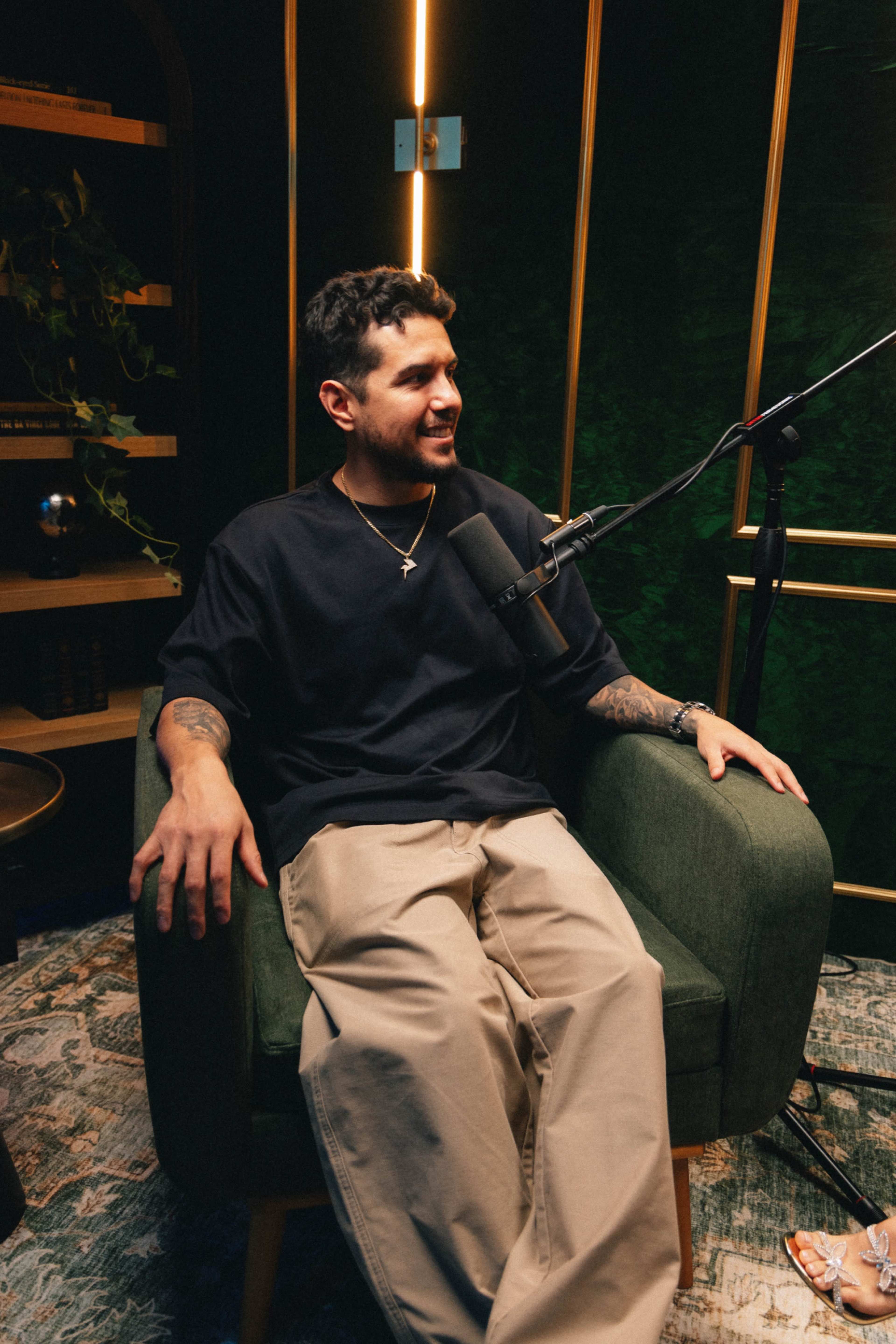 A man sits in a green armchair in a well-lit studio, speaking into a microphone.
