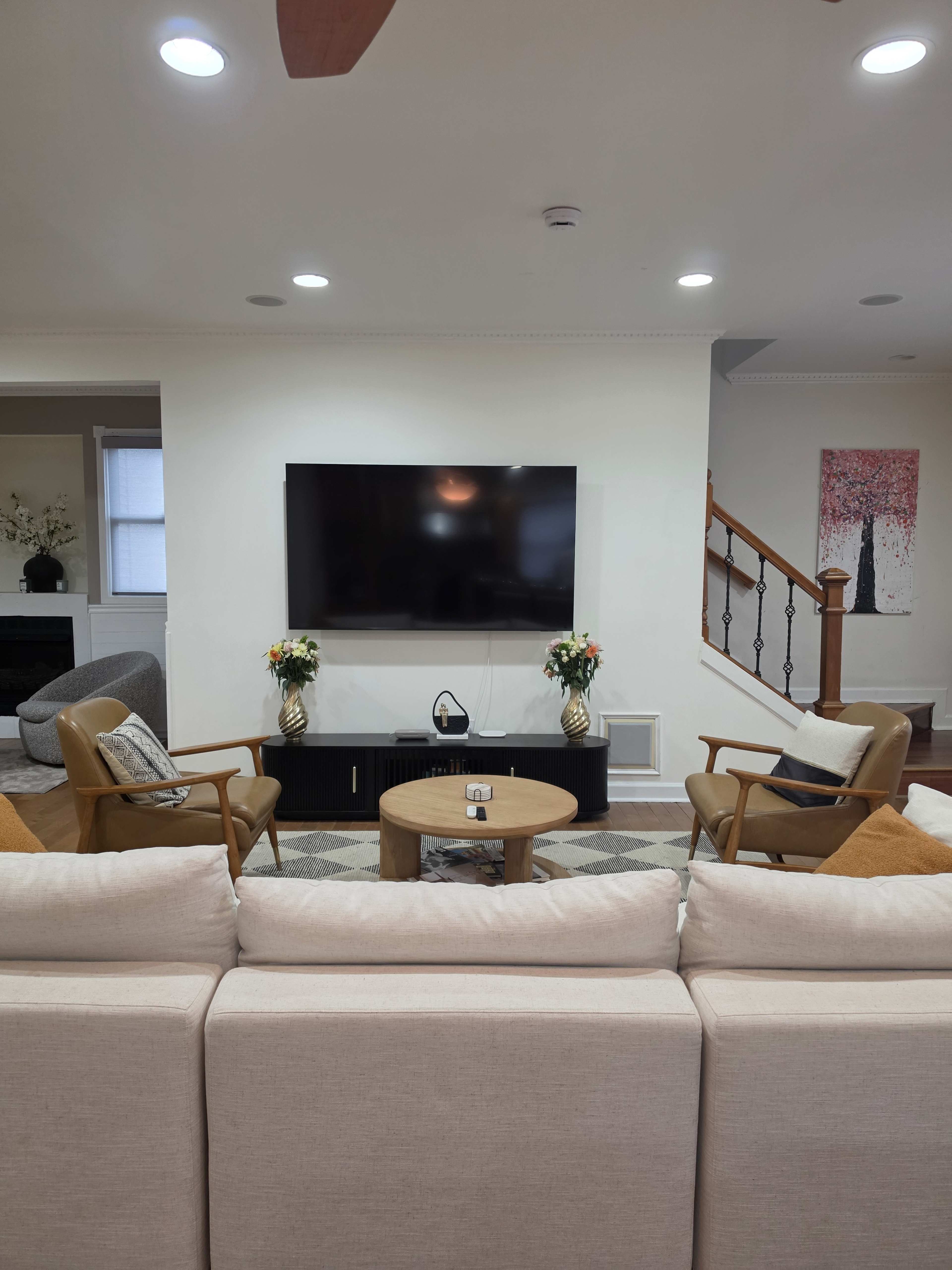 The image shows a modern living room with a large flat-screen TV mounted on the wall, two armchairs, and a coffee table in front of a beige sectional sofa.