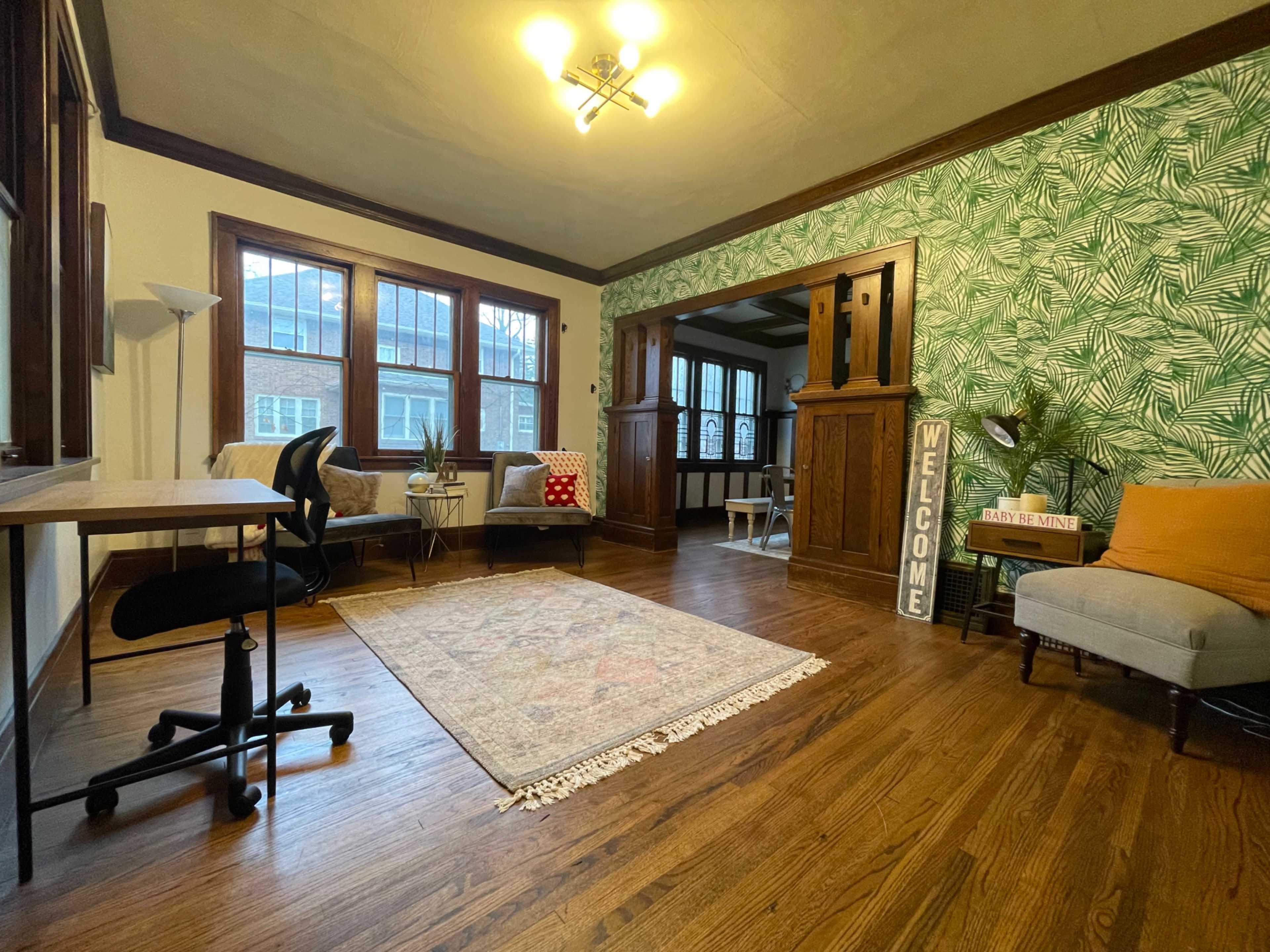 A living room with hardwood floors, a patterned area rug, a desk and chair, and tropical-themed wallpaper.