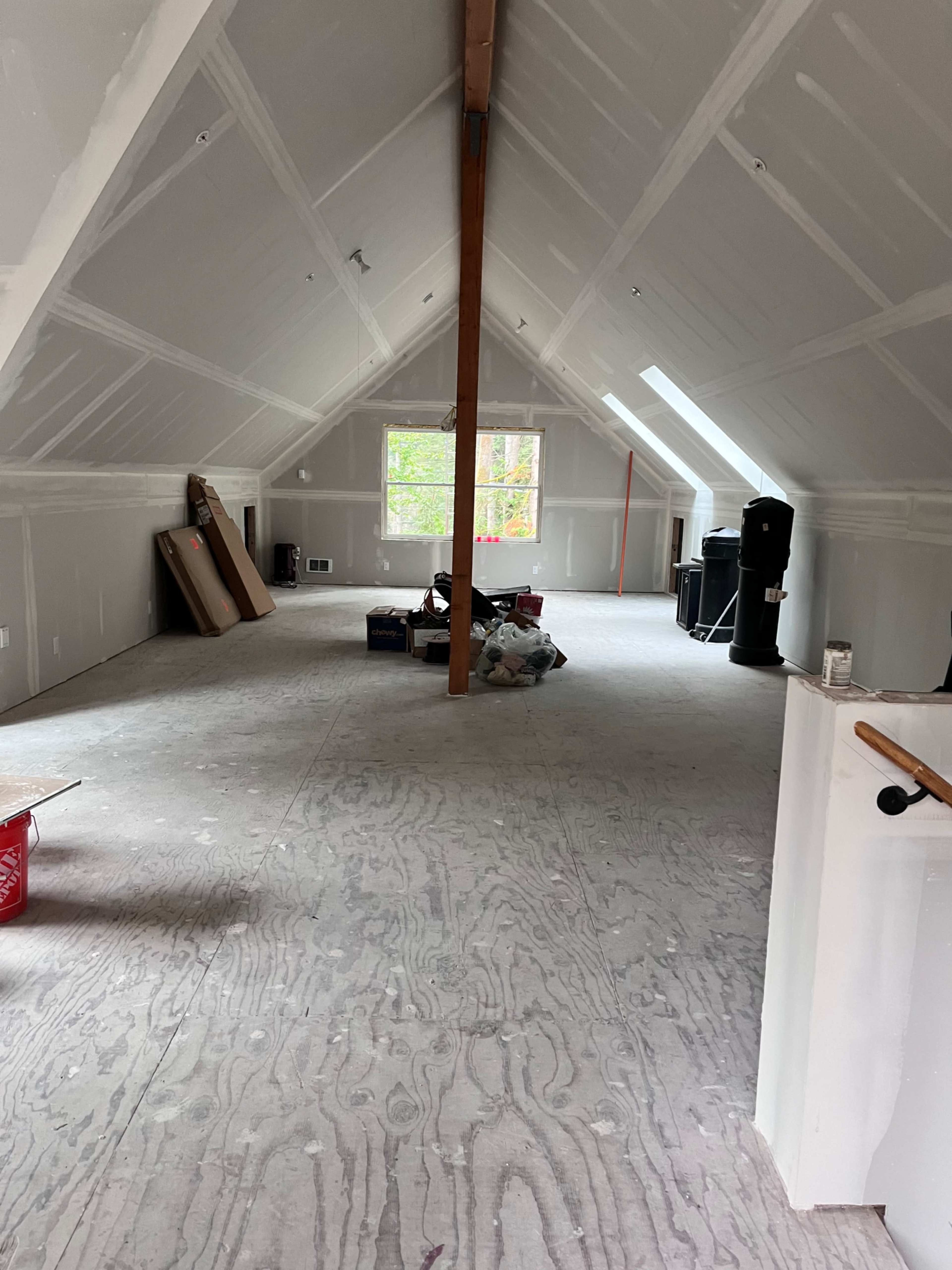 The image shows a spacious, unfinished attic with exposed beams, a large window, and construction materials scattered across the floor.