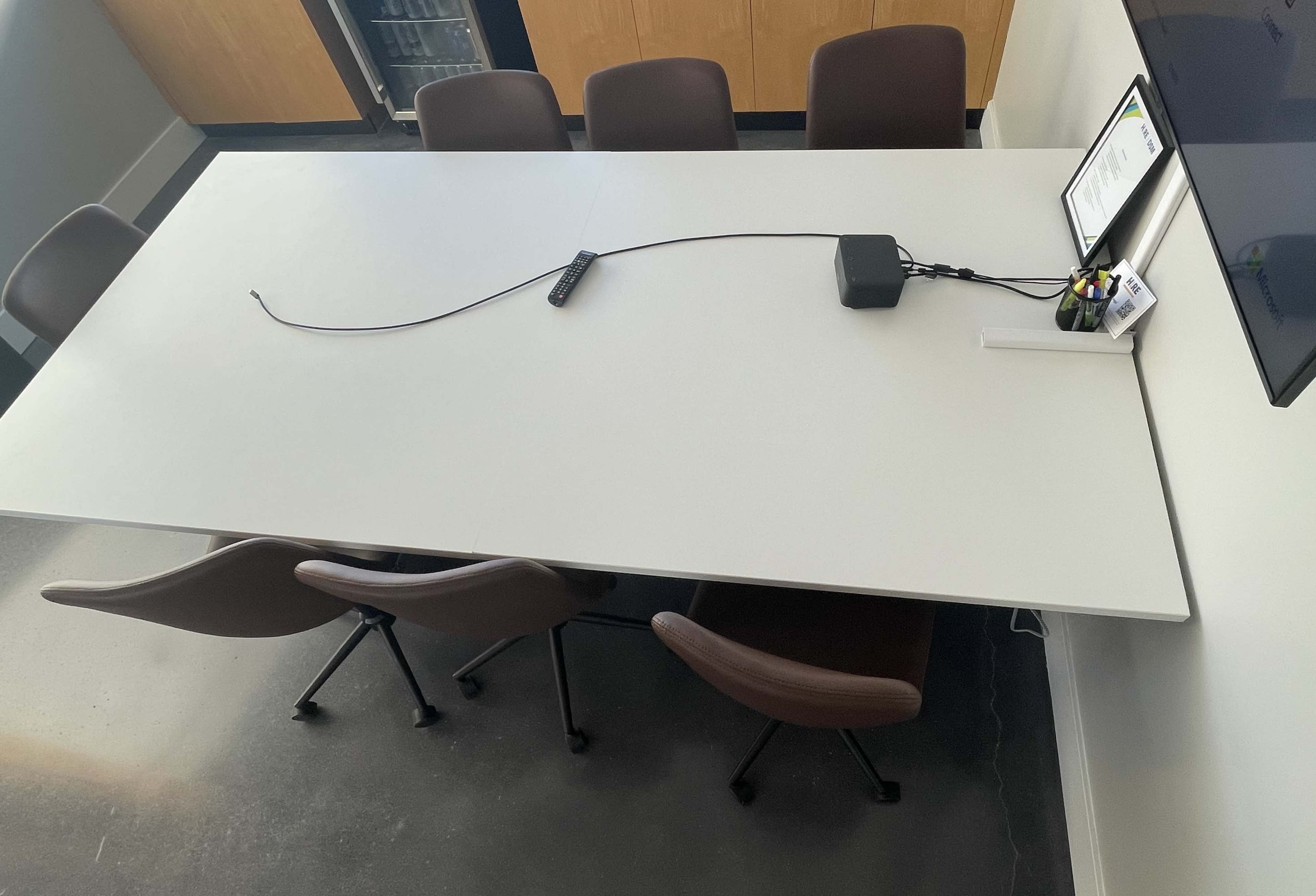 A large conference table surrounded by several brown chairs, with a cable connecting to a device on the table's surface, against a background of cabinets and a mounted screen.