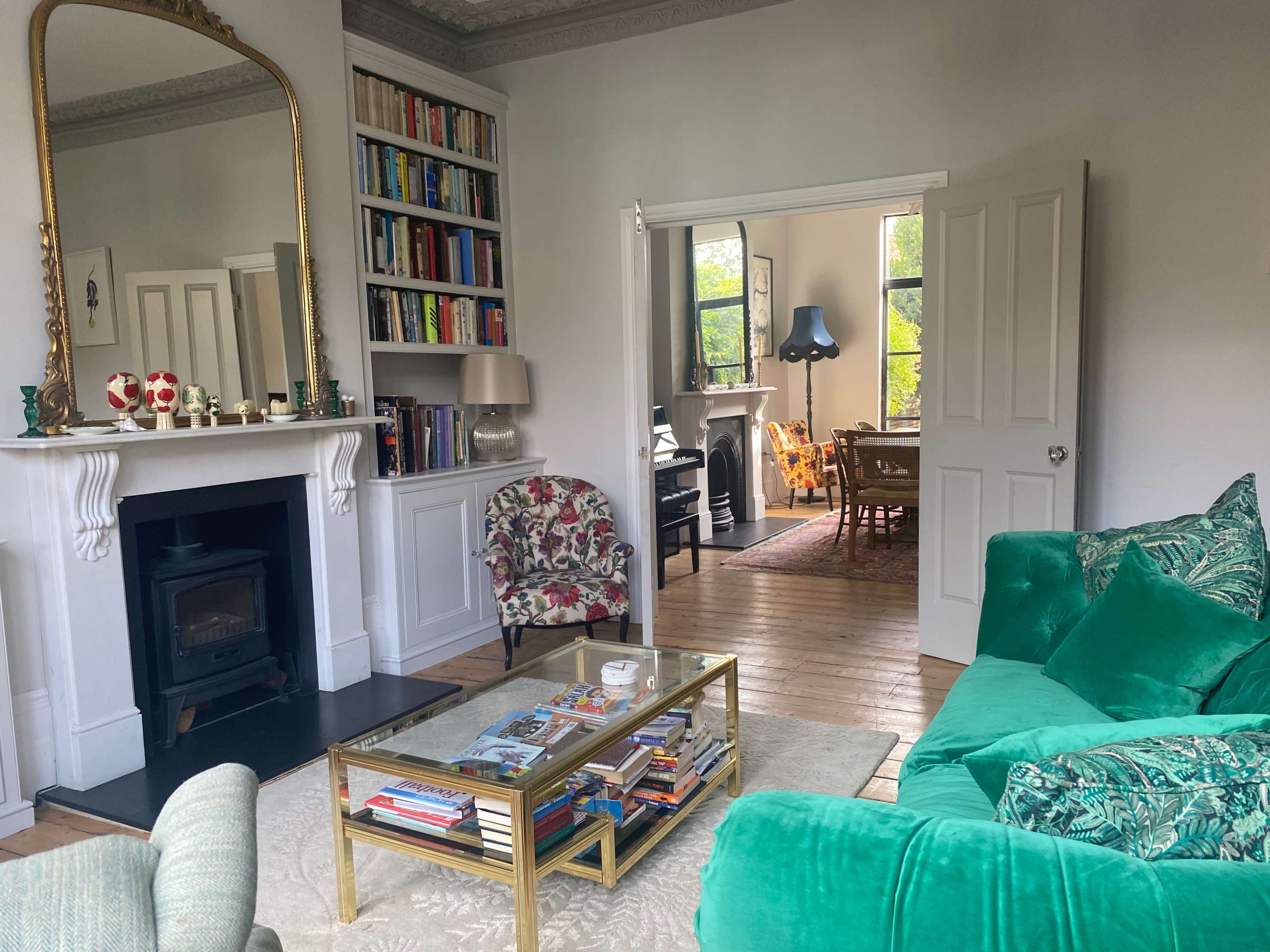 A cozy living room with a green sofa, a coffee table stacked with magazines, a bookshelf, and a view into a well-lit dining area.