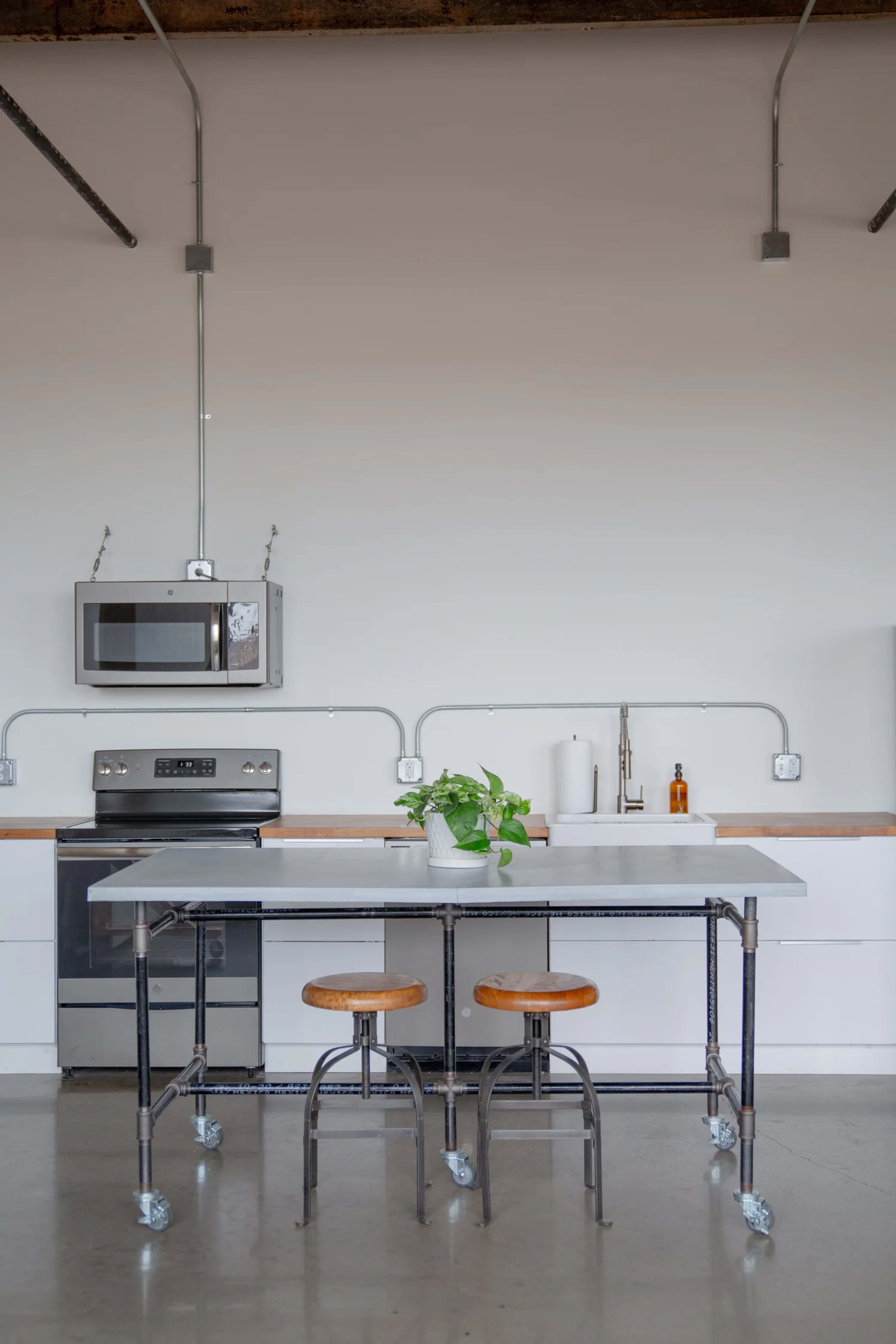 The image shows a modern kitchen with a stainless steel countertop, two wooden stools, and essential appliances including a microwave and an oven, set against a plain white wall.