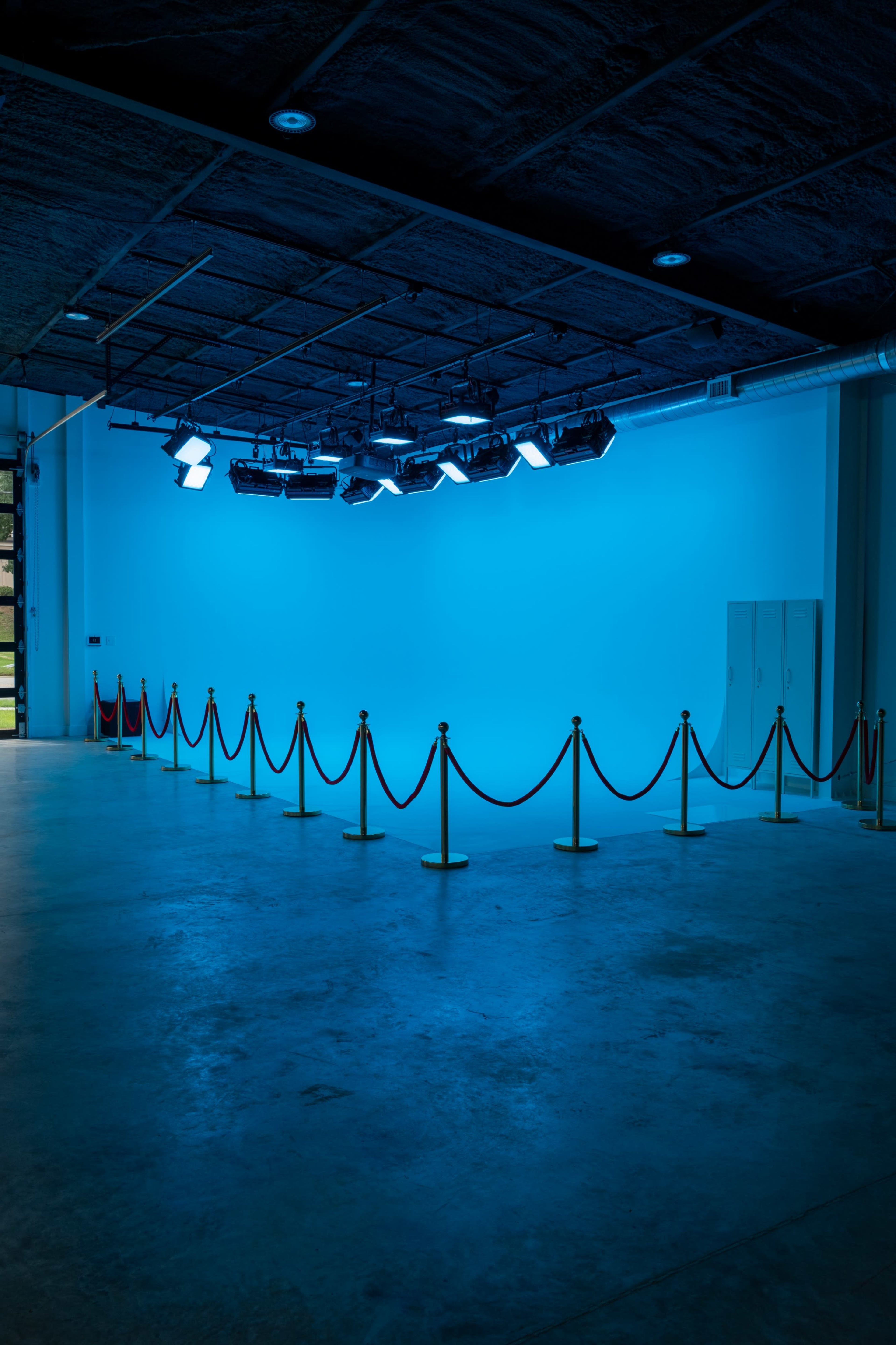 The image shows a spacious, empty area with blue lighting, marked by a stanchion rope barrier in front of a plain wall.