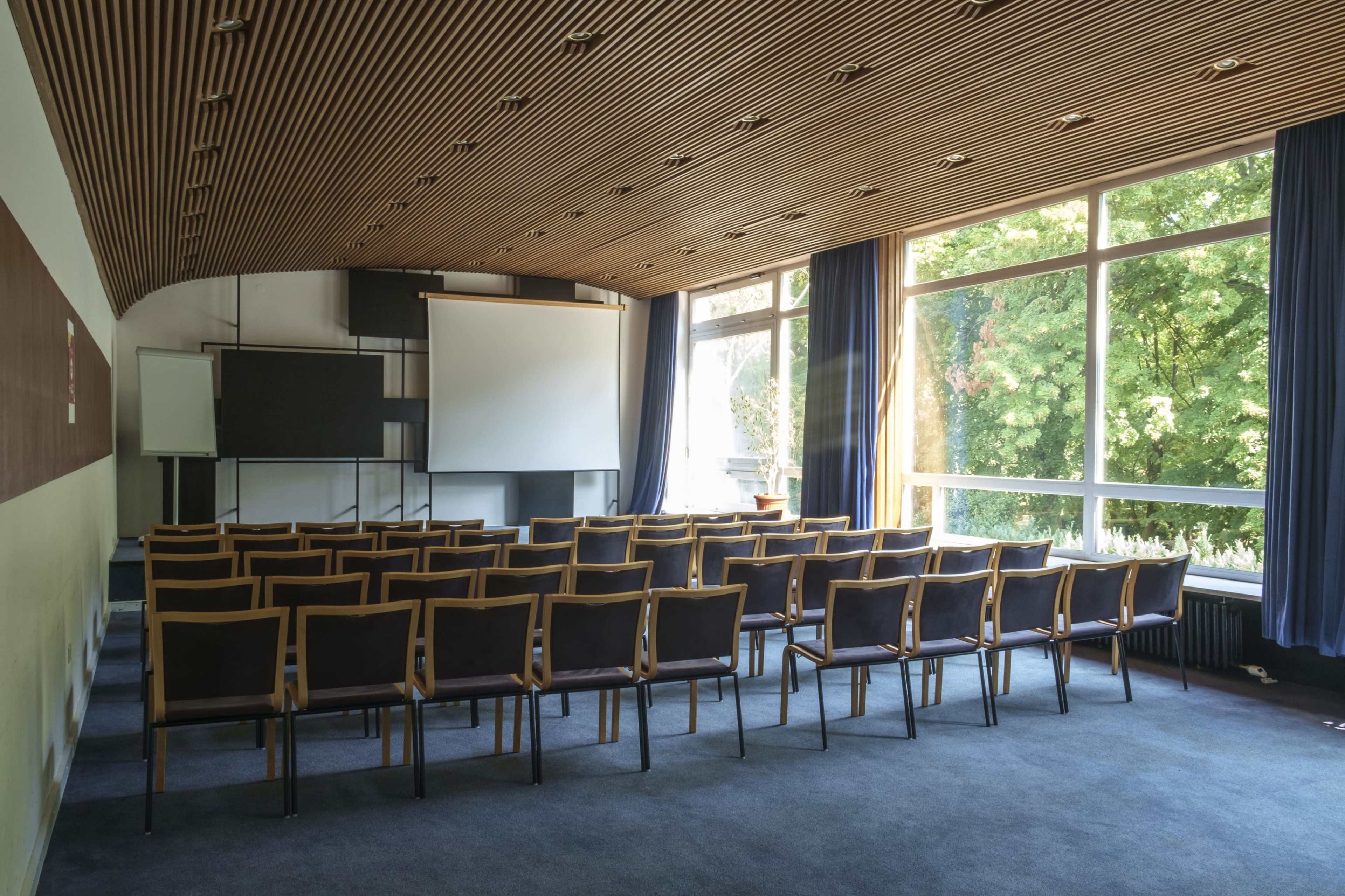 The image shows a sparsely furnished conference room with rows of chairs facing a large projection screen and large windows overlooking greenery.