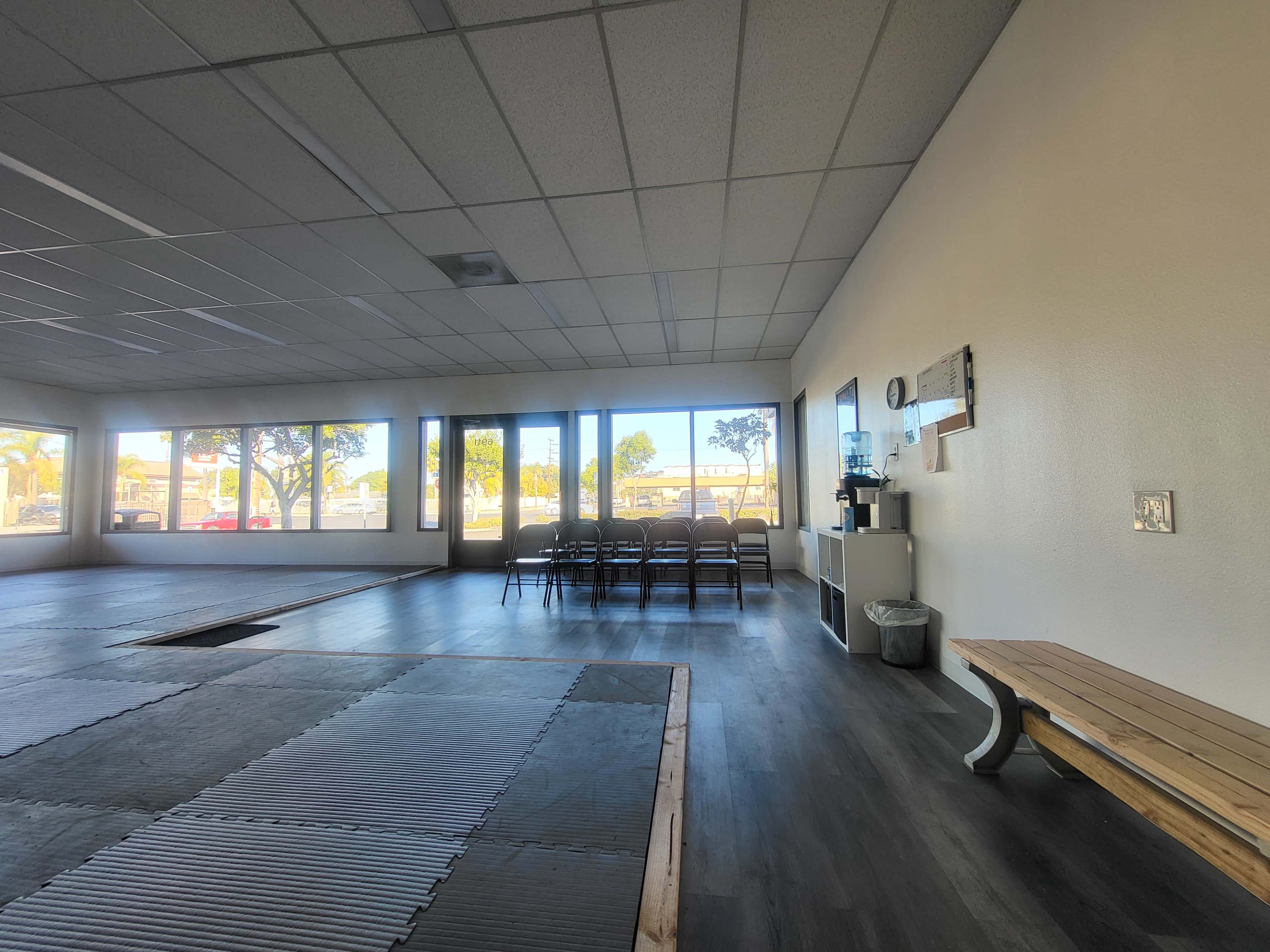 The image shows a spacious training room with wooden flooring, a bench, a water cooler, and several empty chairs arranged against the wall, illuminated by natural light from large windows.