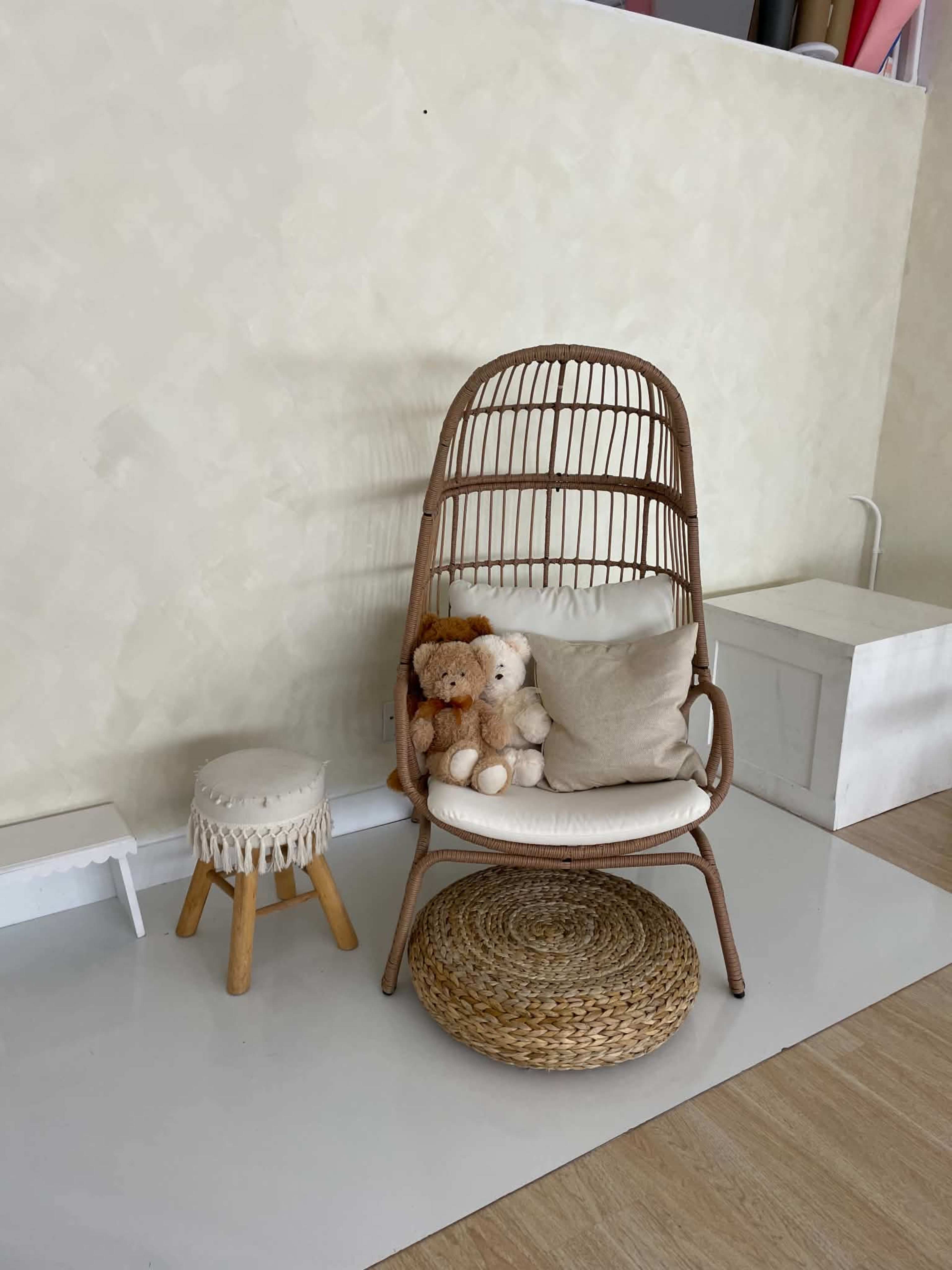 A rattan chair with cushions and stuffed animals is positioned next to a small wooden stool on a light wooden floor.
