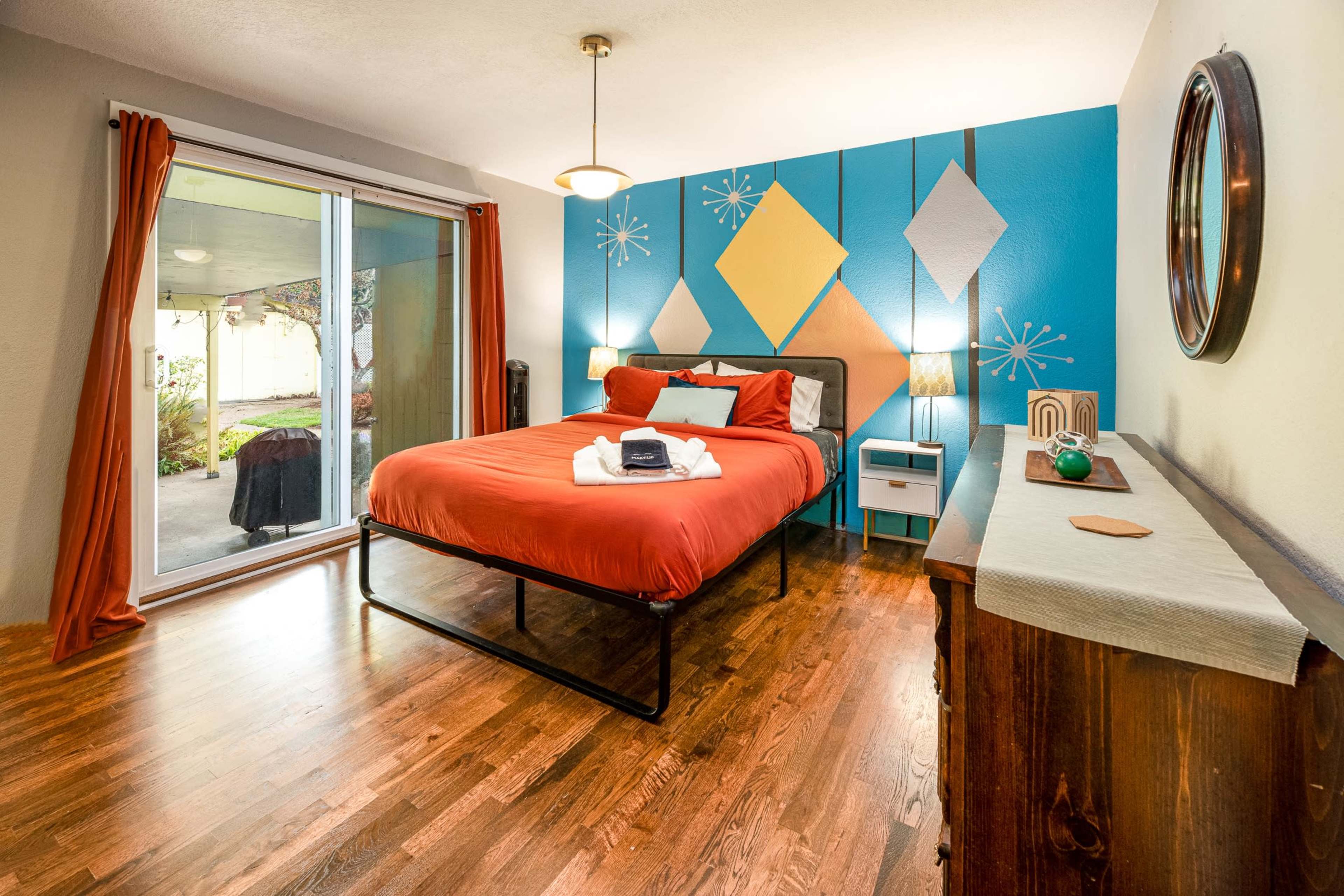 The image shows a neatly arranged bedroom with a bed featuring an orange duvet, a sliding glass door leading to an outdoor space, and colorful geometric wall art.