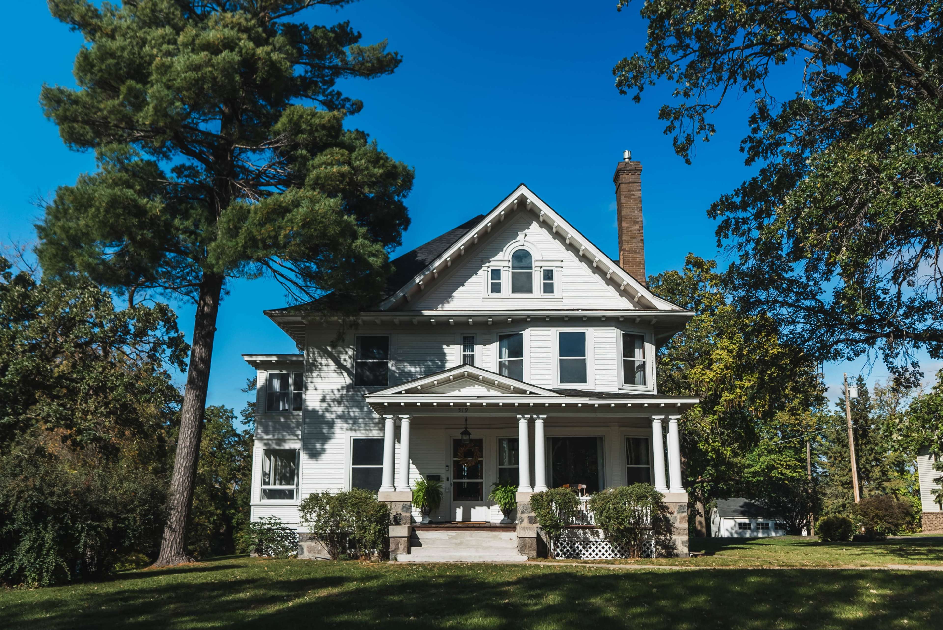A large, two-story white house with a porch and tall trees stands on a grassy lawn under a clear blue sky.