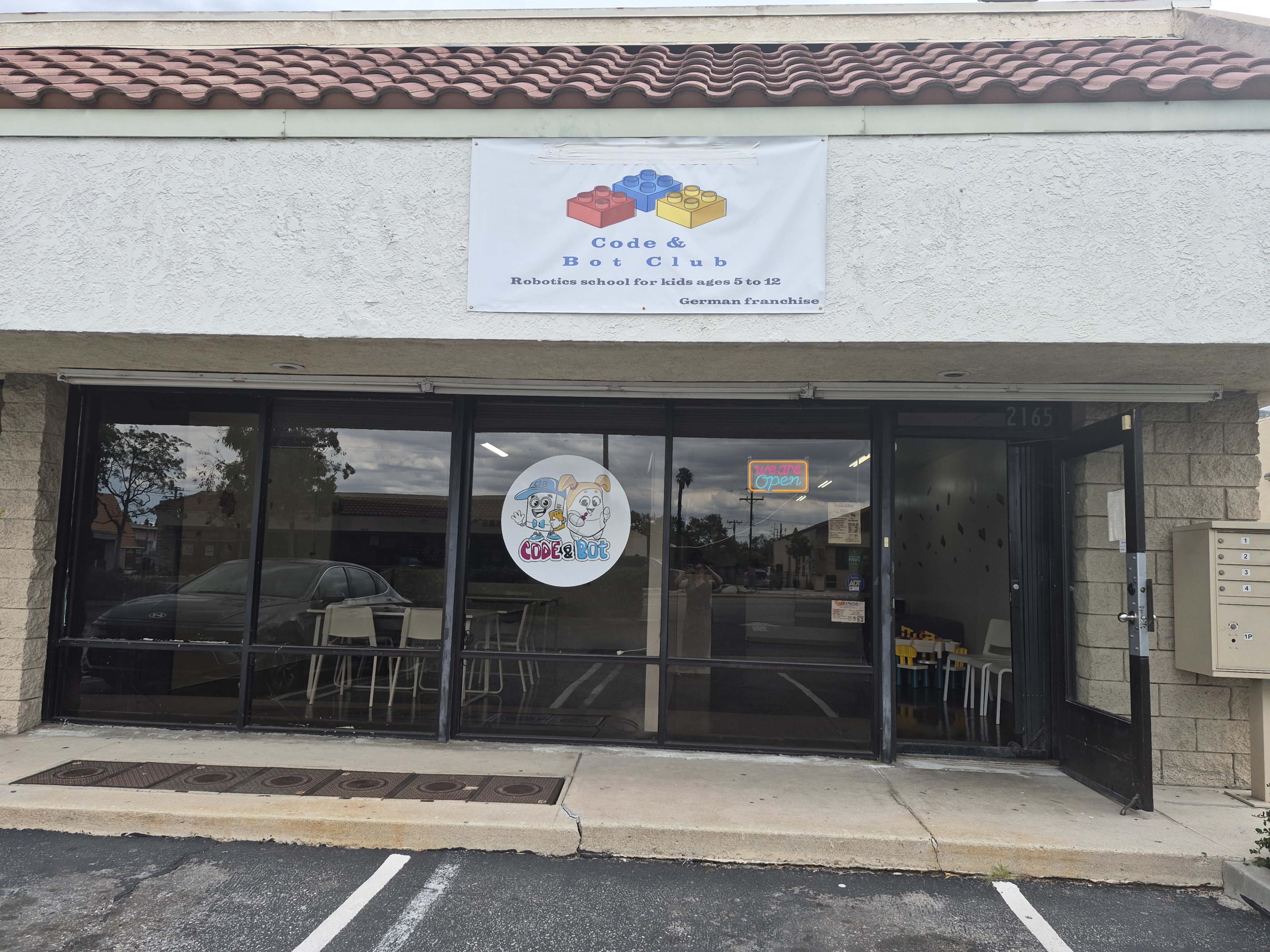 The image shows a storefront for a robotics school called "Code & Bot Club," featuring a colorful sign and a large window displaying playful graphics.