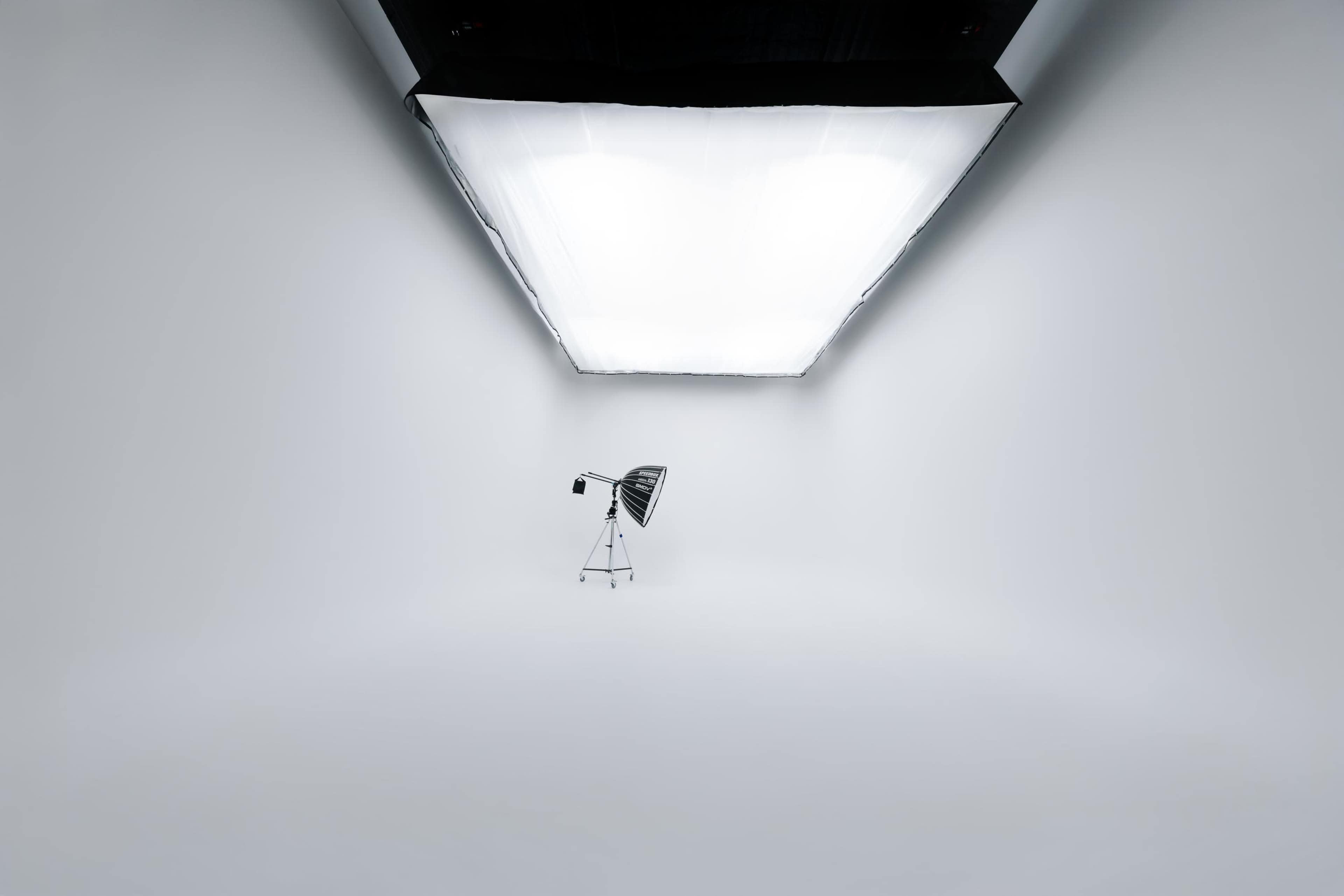 The image shows an empty studio space with a large softbox light mounted on the ceiling and a standing fan positioned in the center.