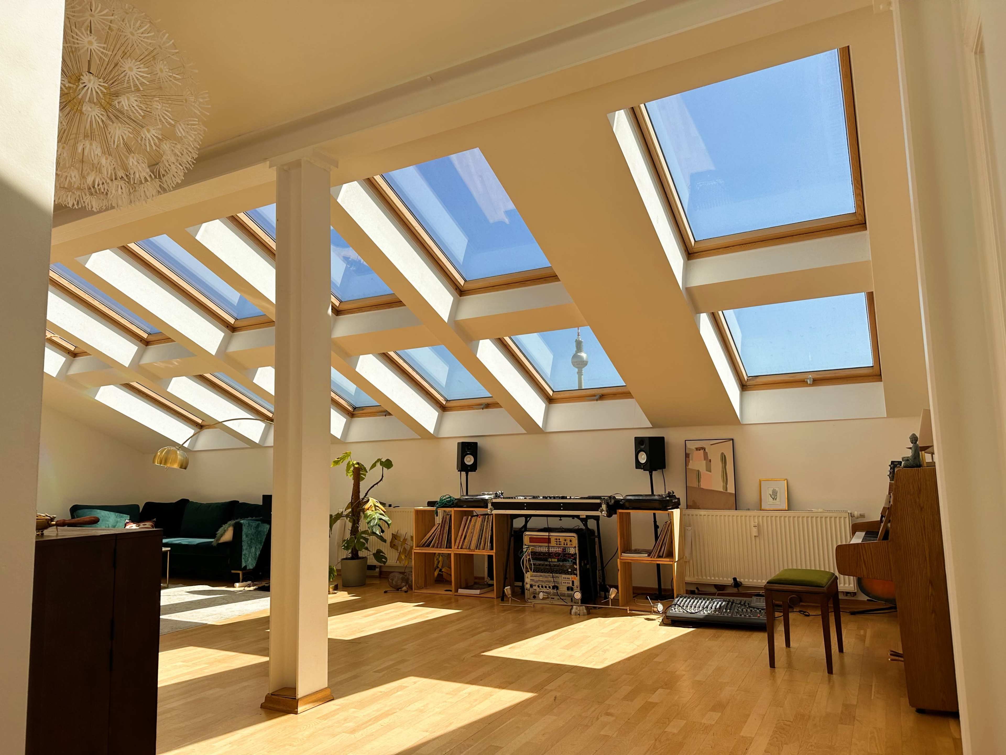 The image shows a spacious, well-lit room with large skylights, wooden flooring, and a music setup along one wall.