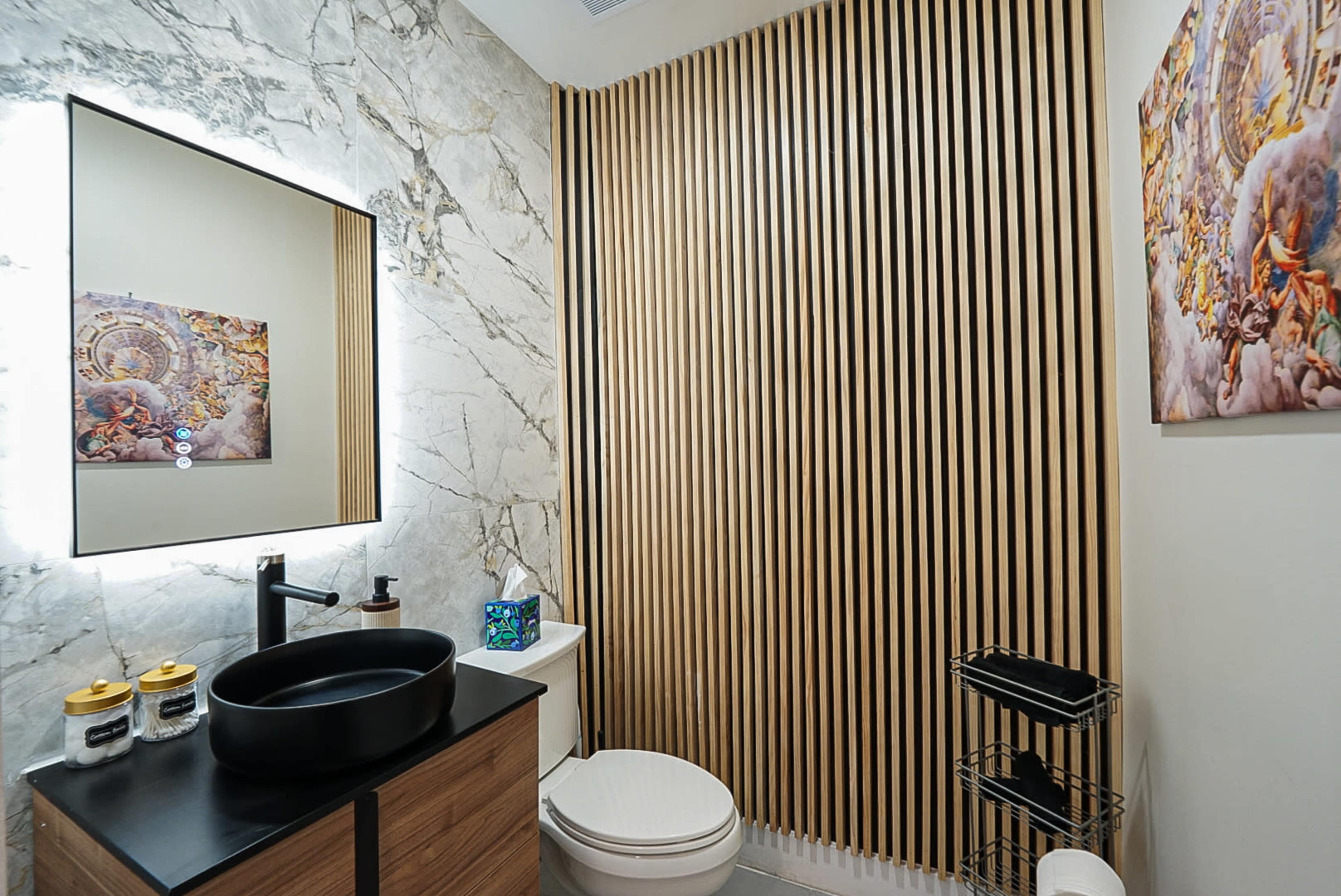 The image shows a modern bathroom featuring a wooden slat wall, a circular mirror, and a black sink against a marble backdrop.