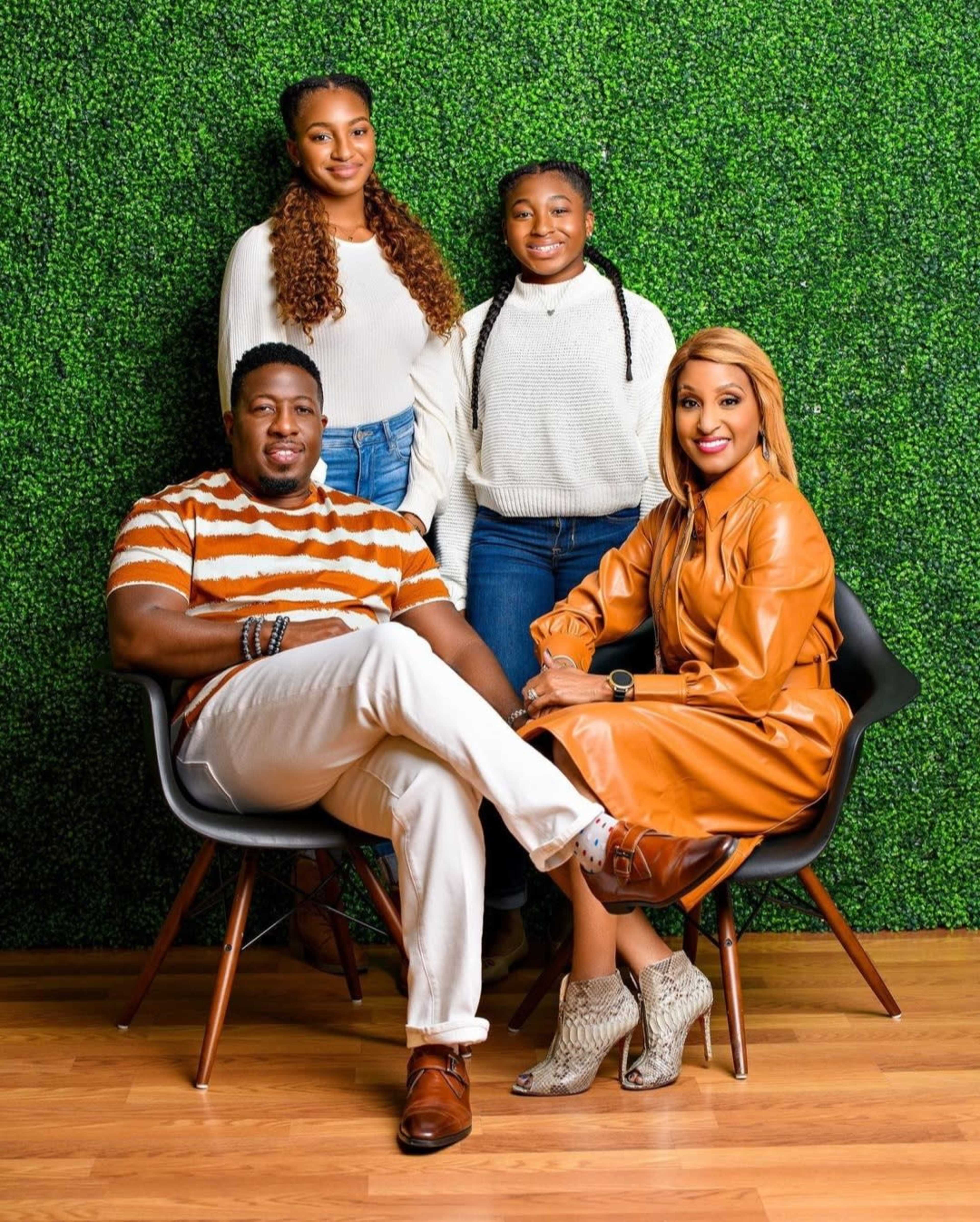 Four people pose for a photo in front of a green artificial hedge wall, with one man seated in a chair and three women standing behind him.