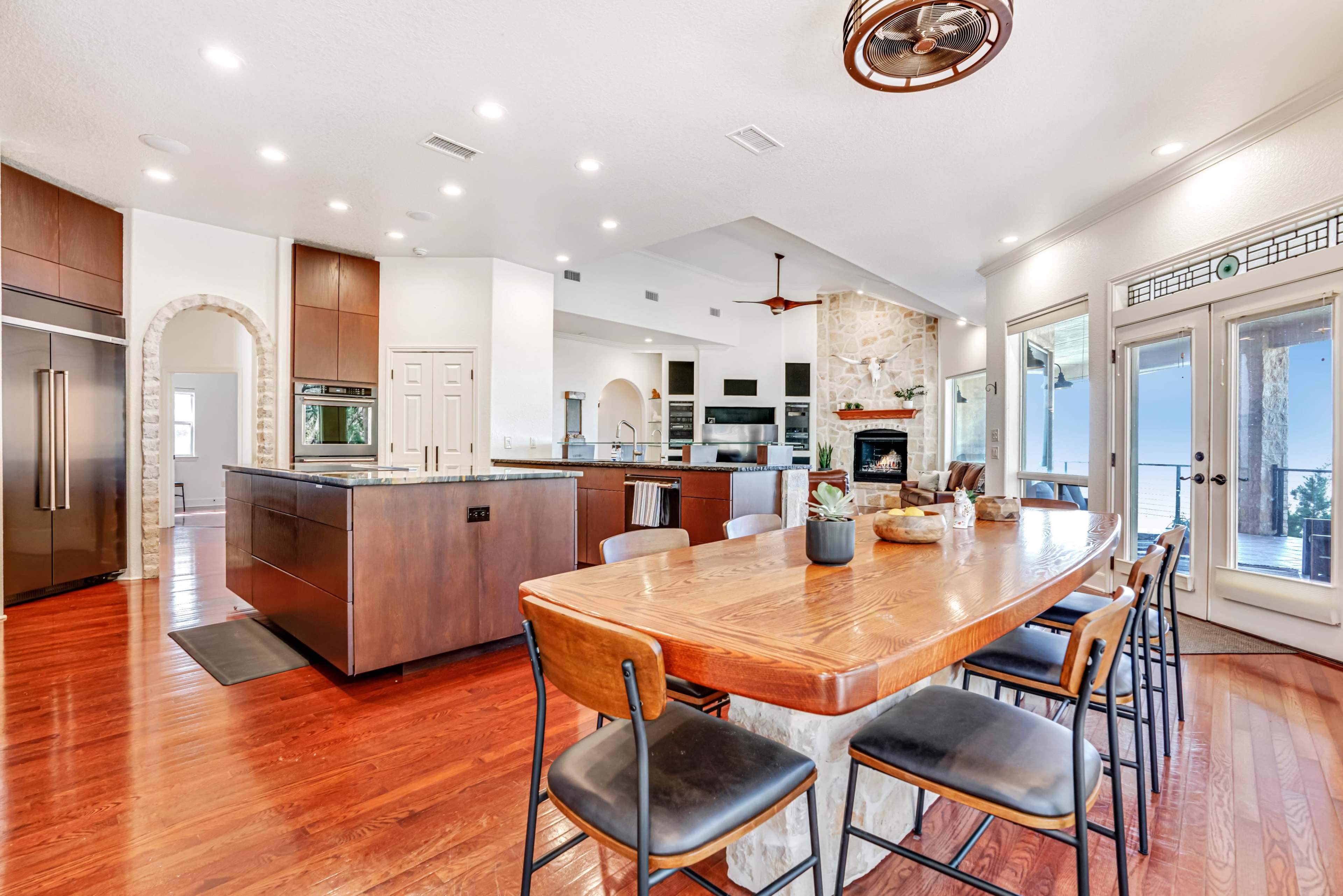 A modern kitchen features a large island with dark cabinetry, a wood dining table, and an open layout leading to a living area with a stone fireplace.