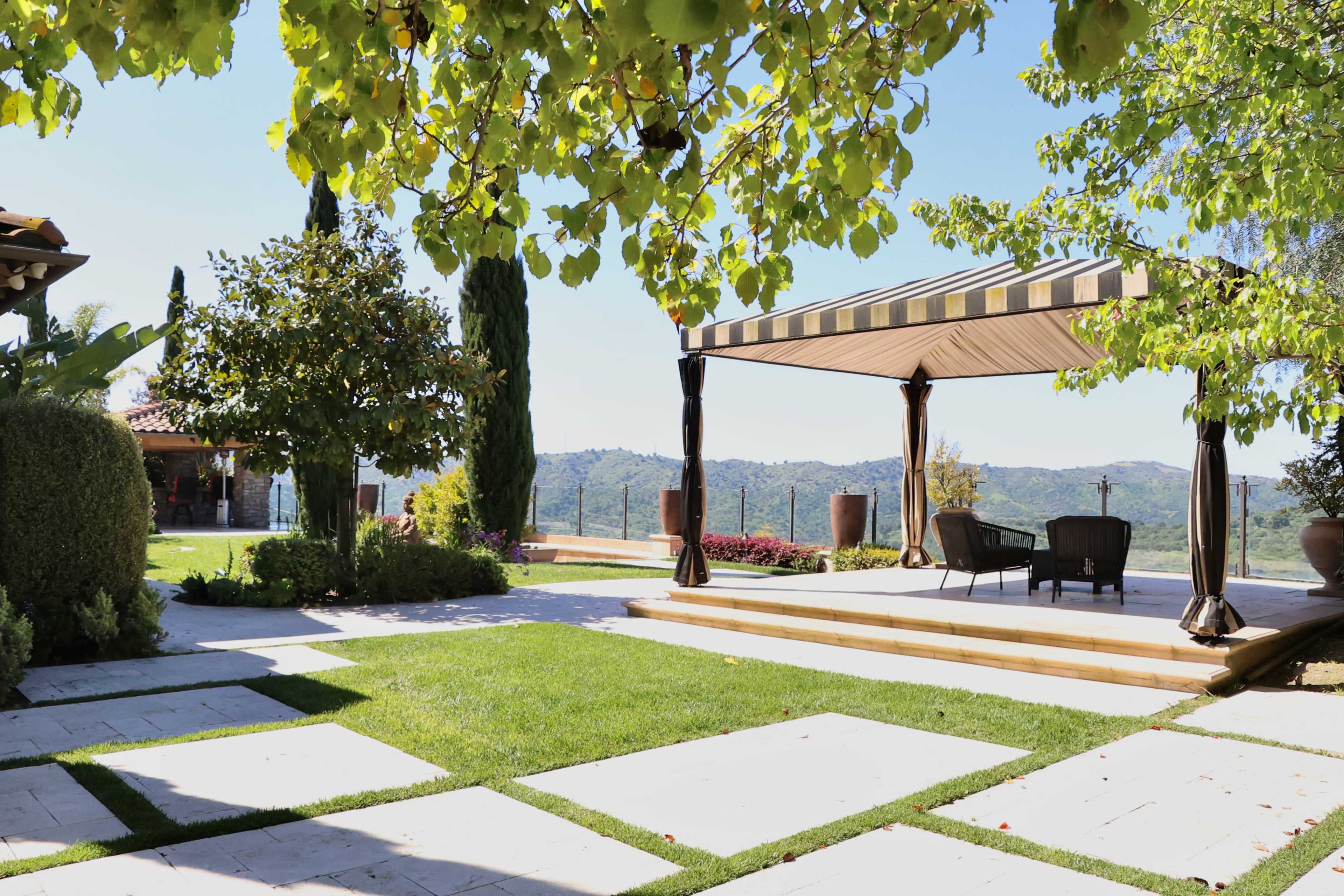 A landscaped outdoor area features stone pathways, a shaded seating space, and views of rolling hills in the background.