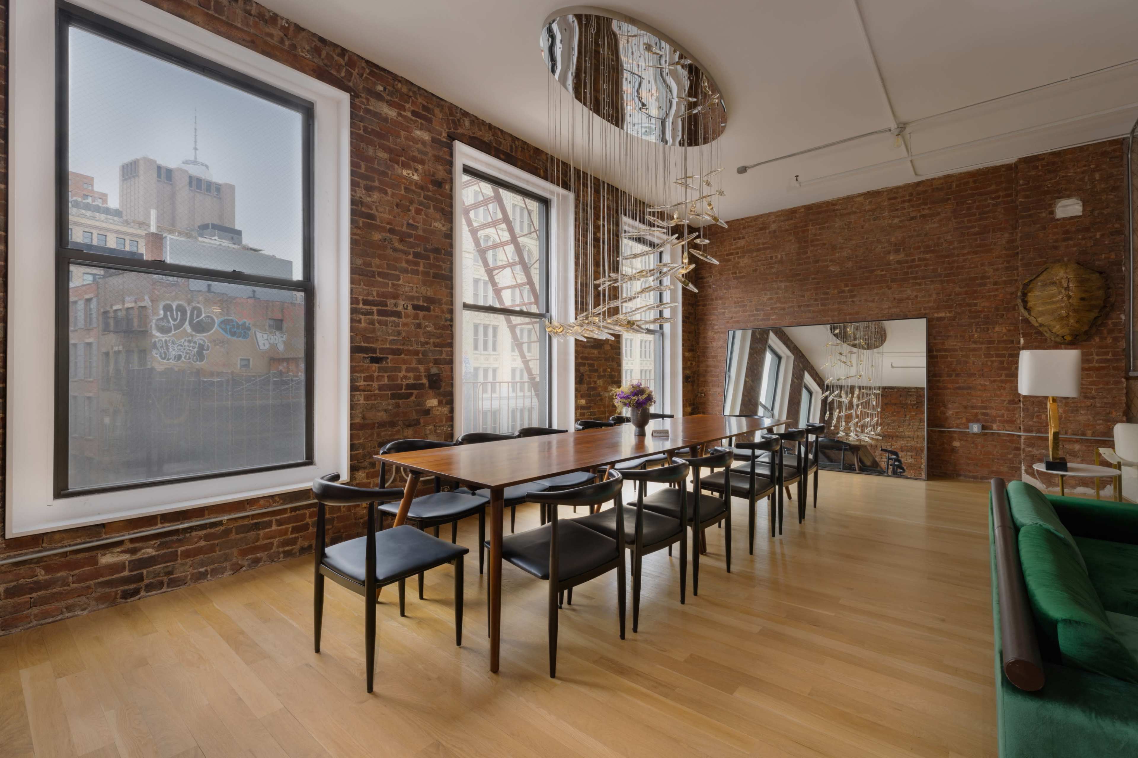 A dining area features a long wooden table surrounded by black chairs, with exposed brick walls and large windows providing city views.