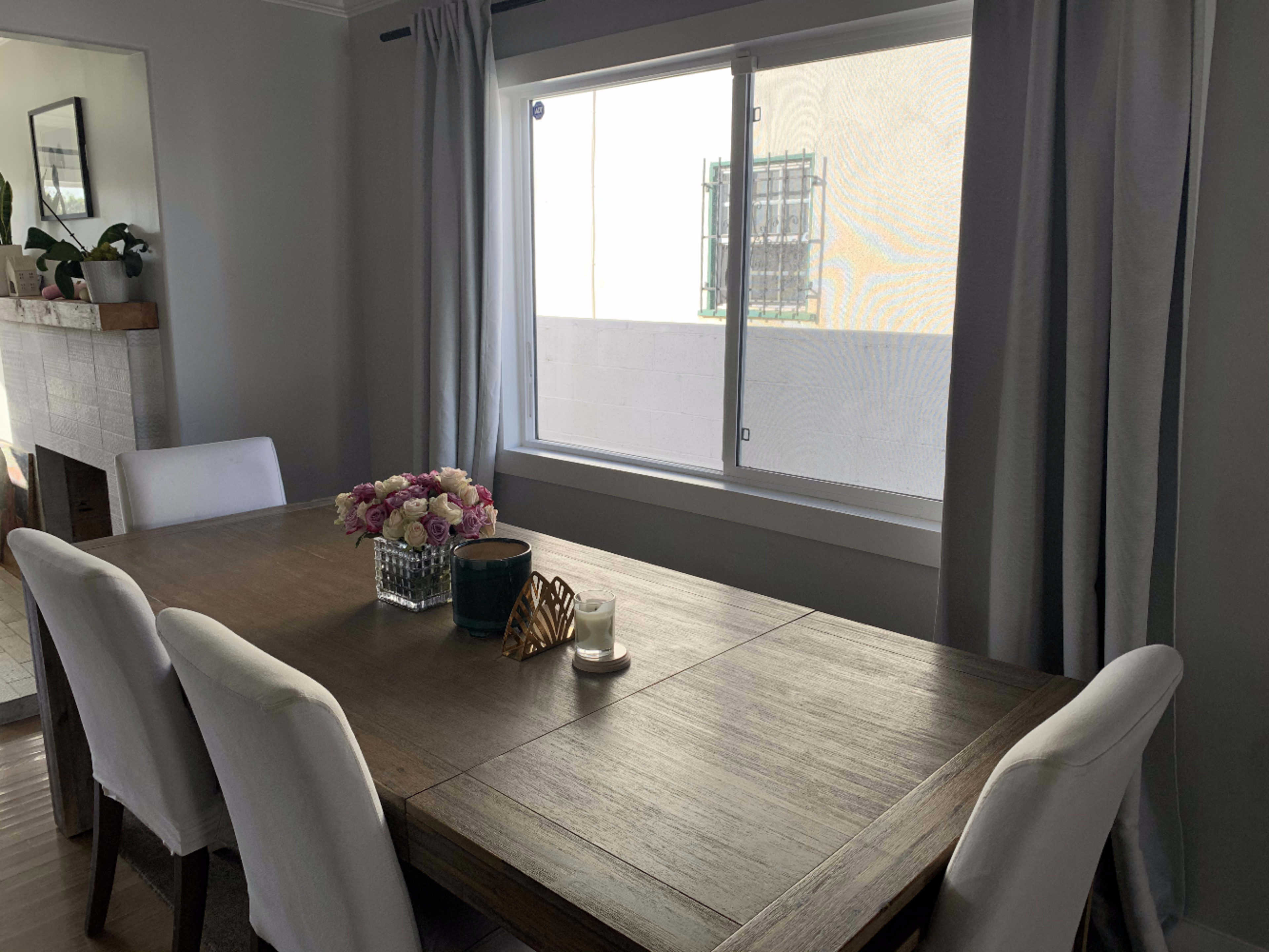 A dining table is set with a vase of flowers in front of a window with sheer curtains.