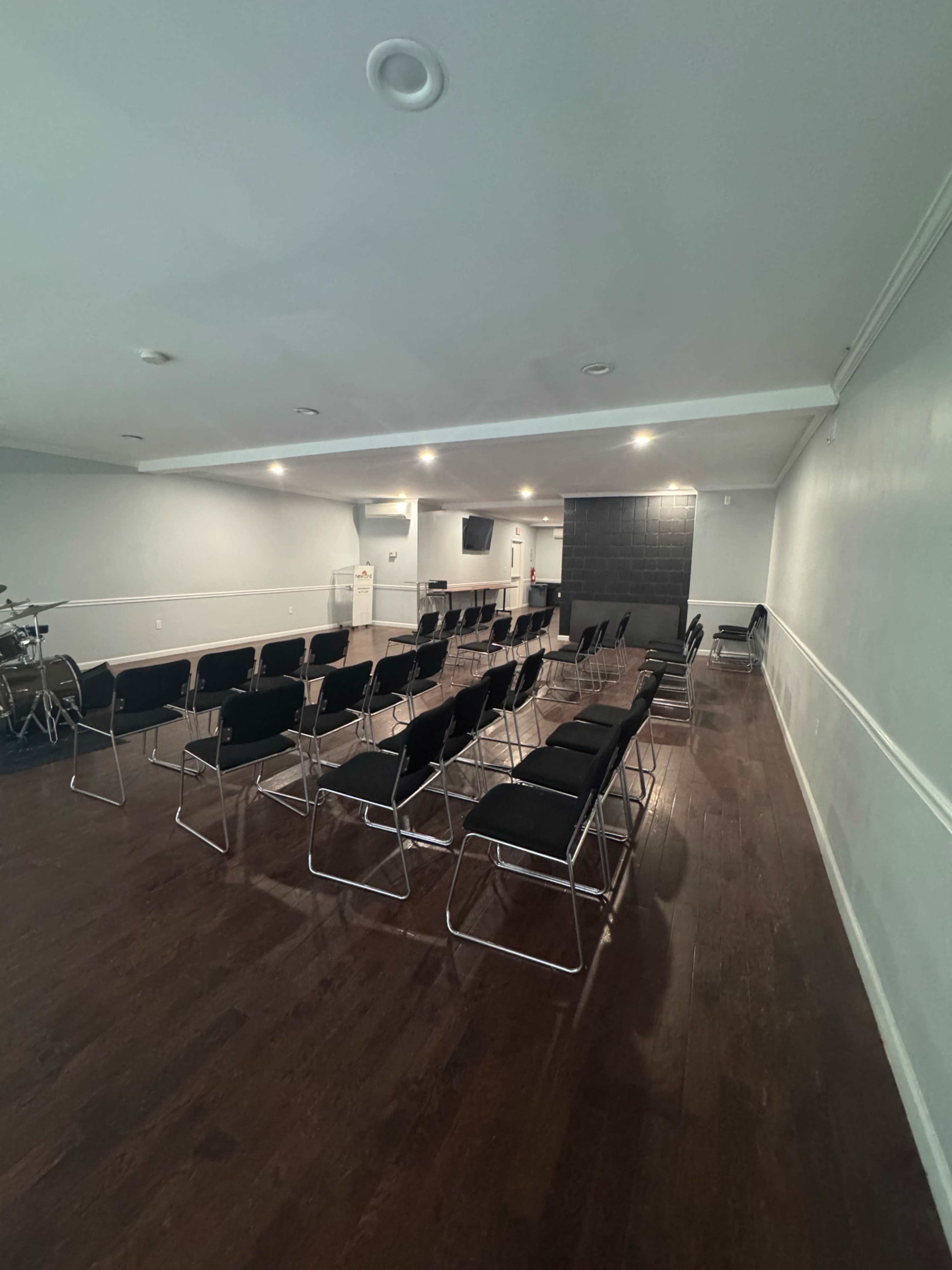 The image shows a large, empty room set up with rows of black chairs facing a wall with a projection screen and sound equipment.