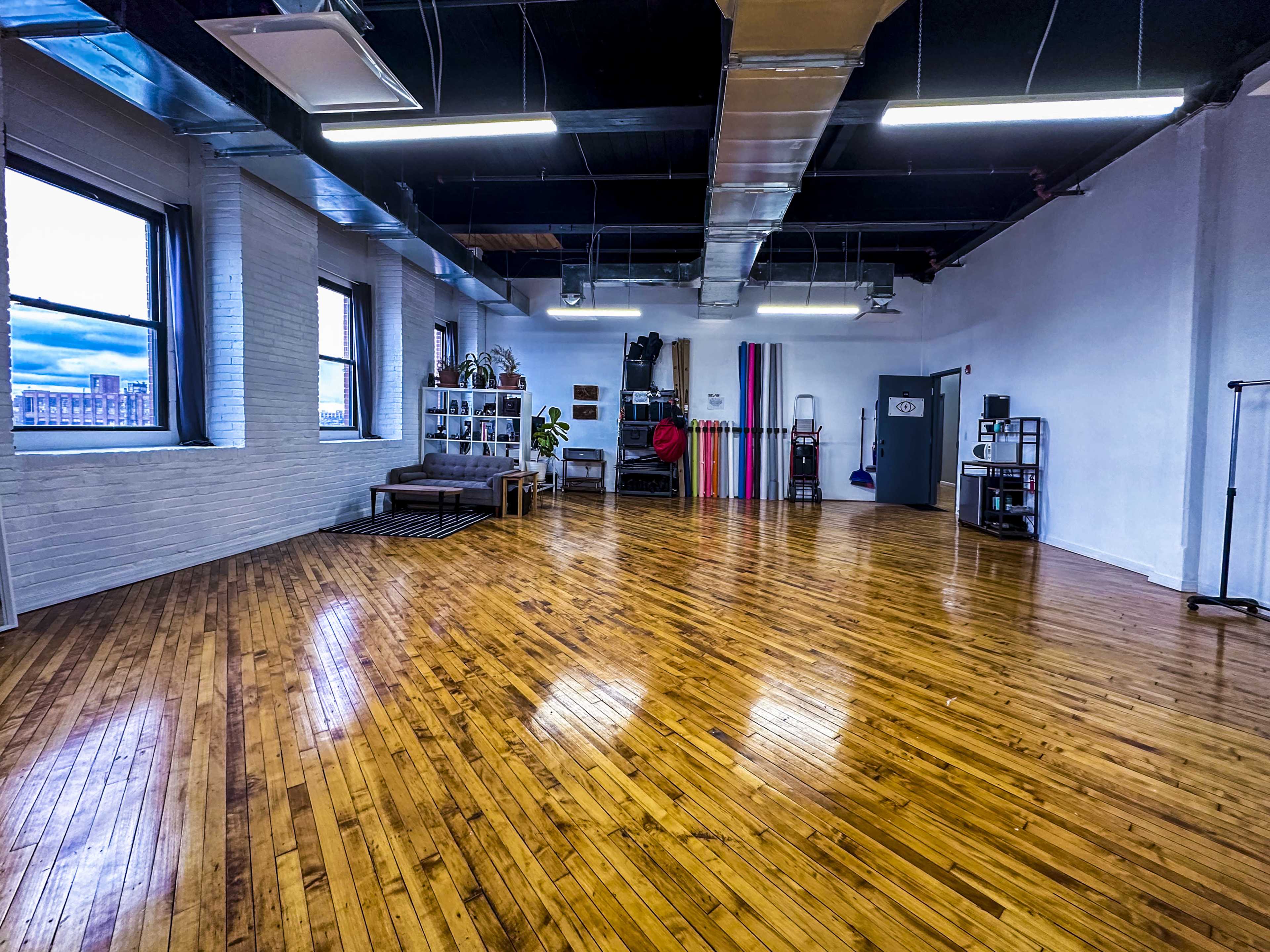 A spacious, brightly lit studio with polished wooden floors, large windows, and various exercise and storage equipment along the walls.