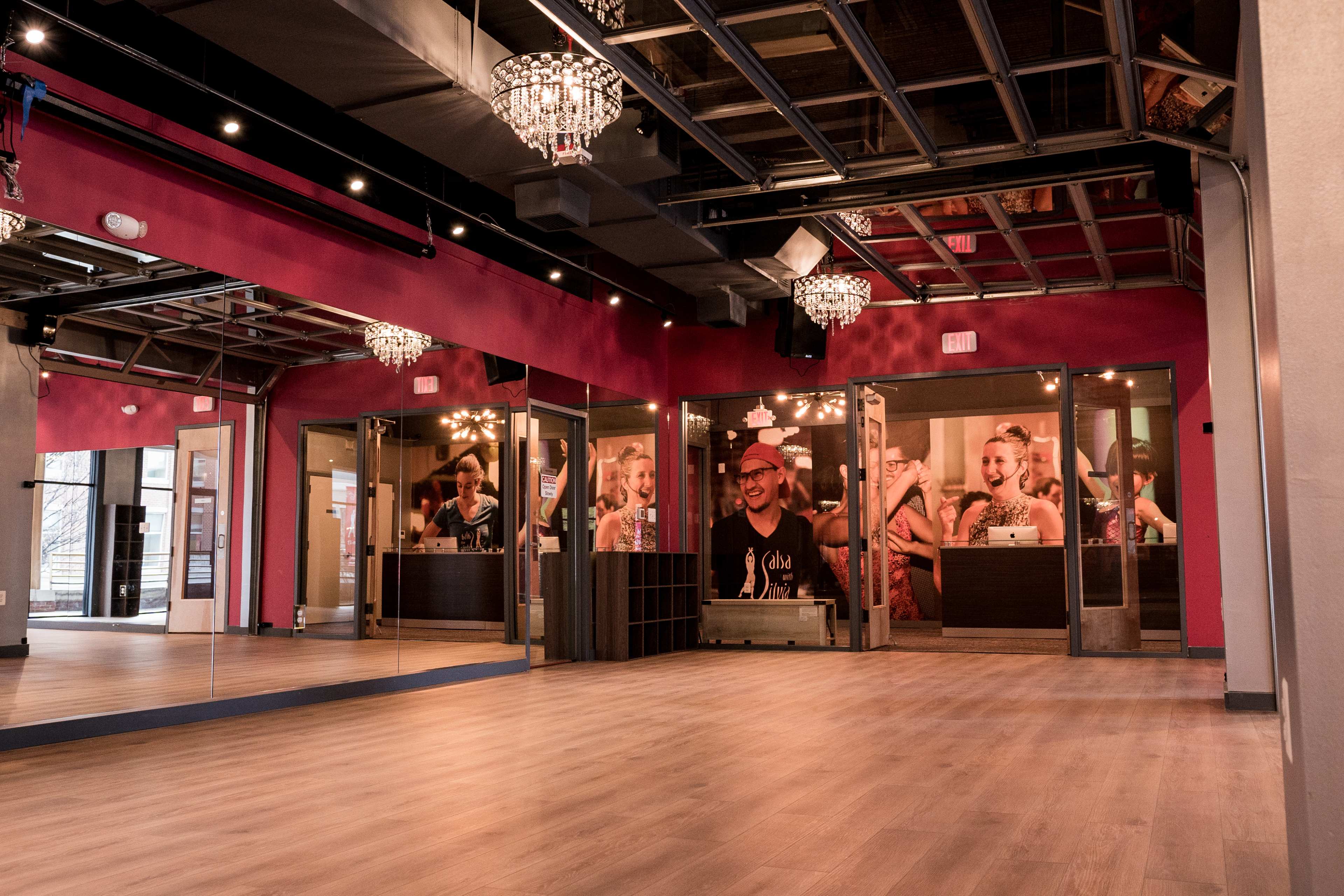 A brightly lit dance studio with wooden floors, large mirrors, and modern lighting fixtures, featuring glass fronted rooms in the background.