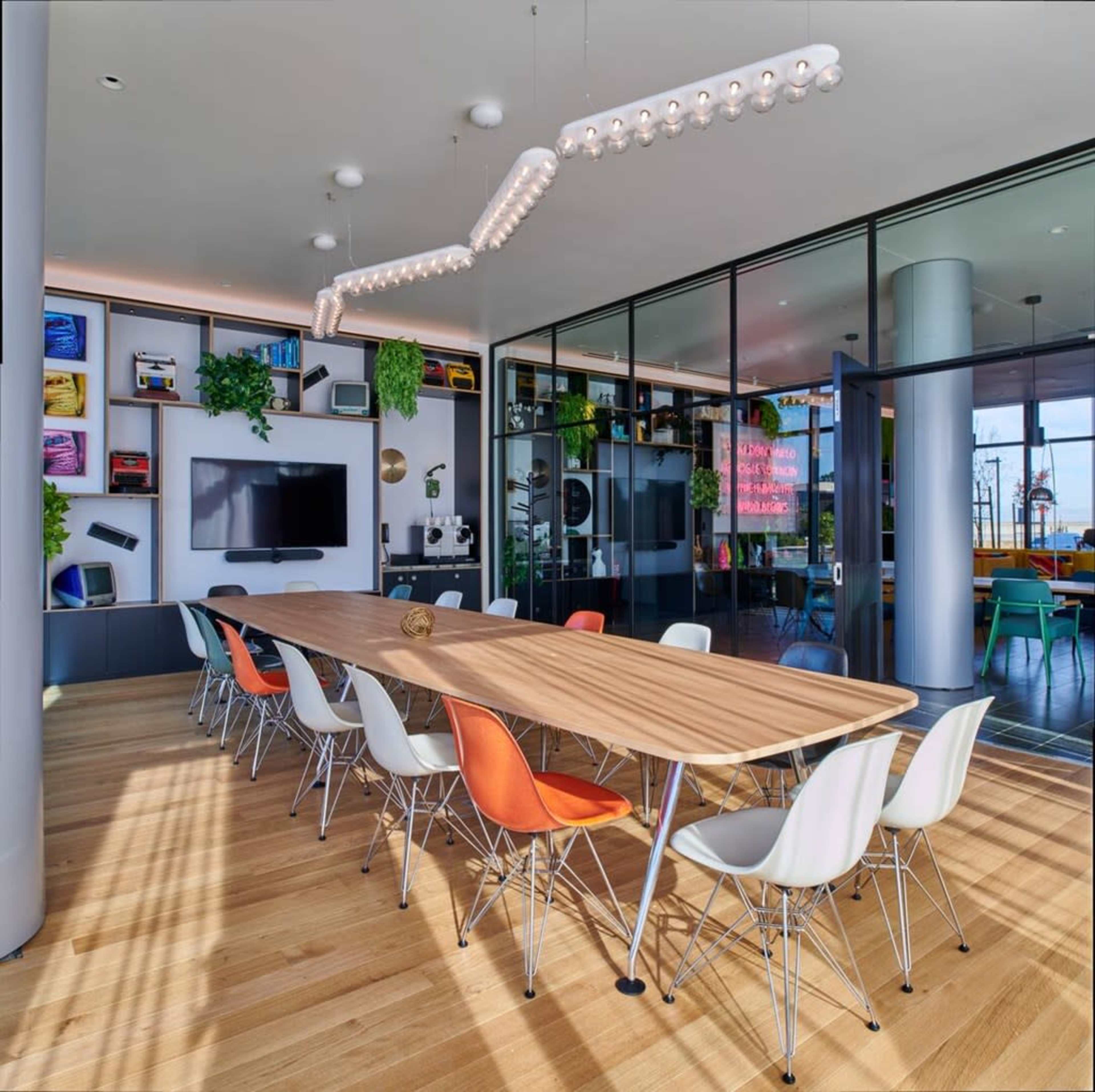 The image shows a modern conference room with a long wooden table surrounded by colorful chairs and large glass walls showcasing a city view.