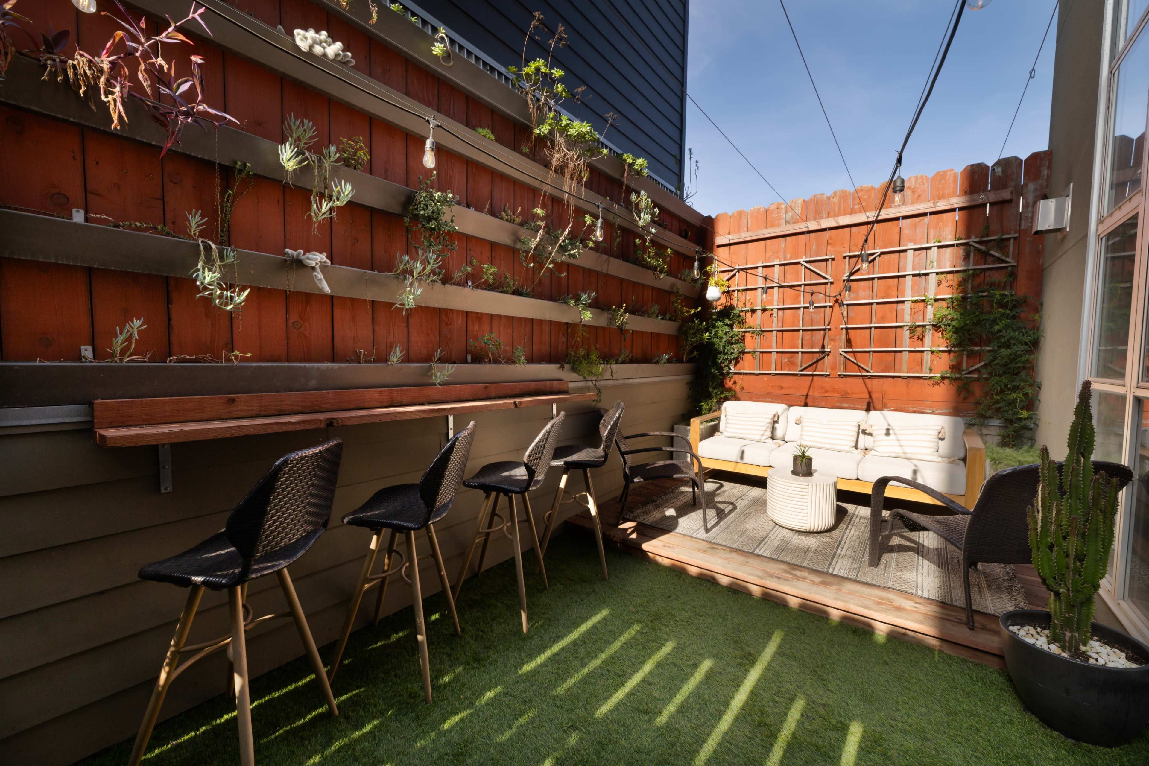 The image shows a small outdoor patio with a wooden fence, a seating area with a white couch and a round coffee table, and several plants along the walls.
