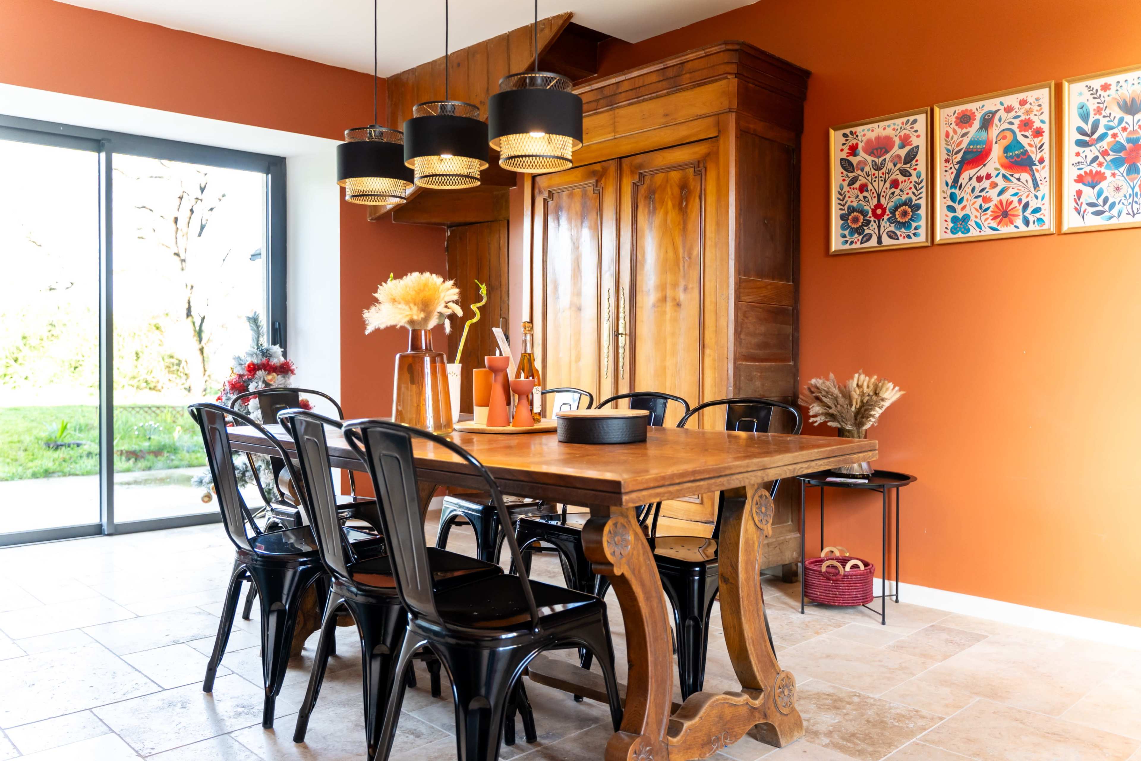 The dining area features a wooden table surrounded by black chairs, with a brown-painted wall and large windows allowing natural light to enter.