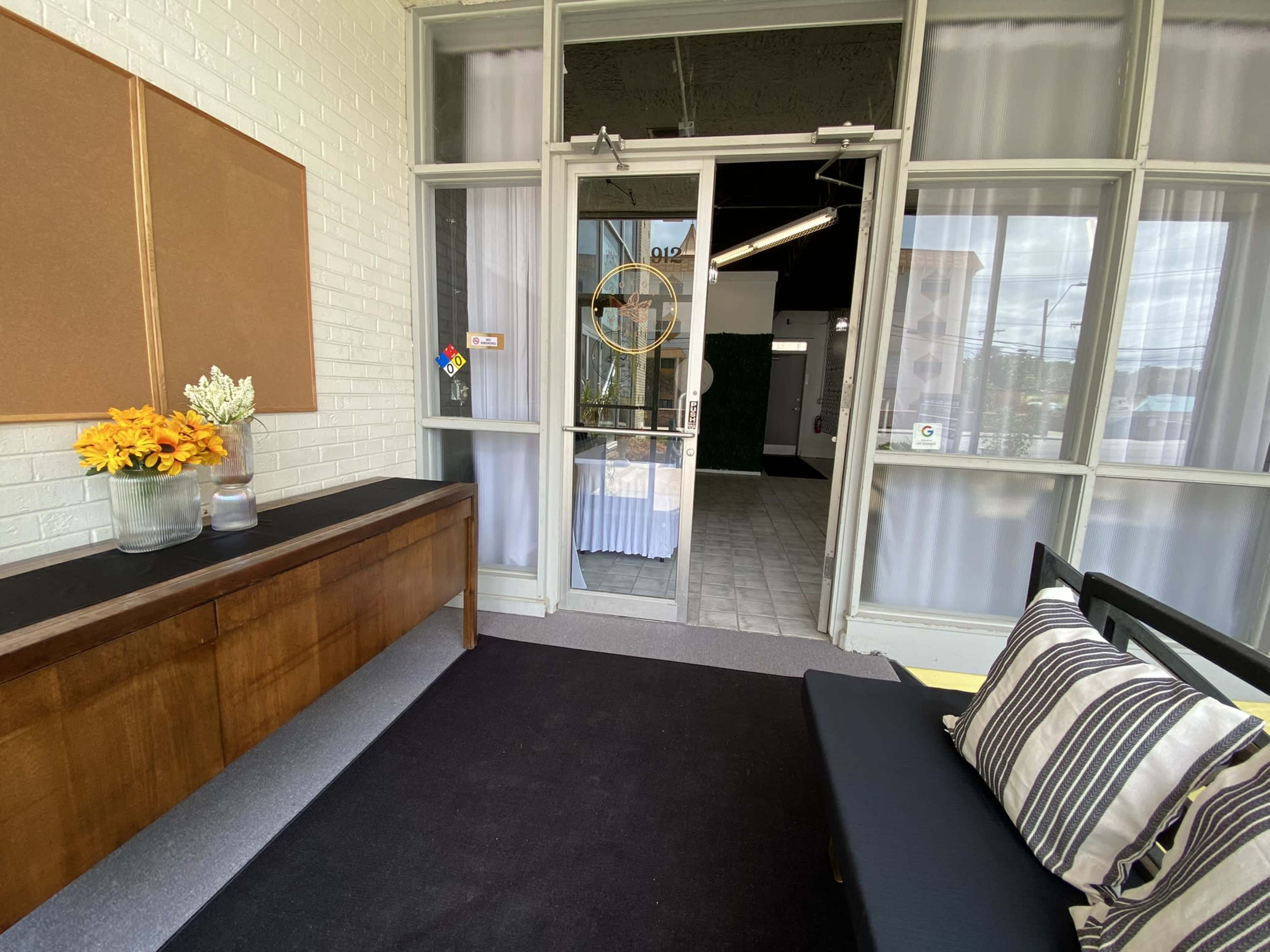 The image shows a well-lit entryway with a low wooden table adorned with flowers, leading to an open door that opens onto a tiled walkway.