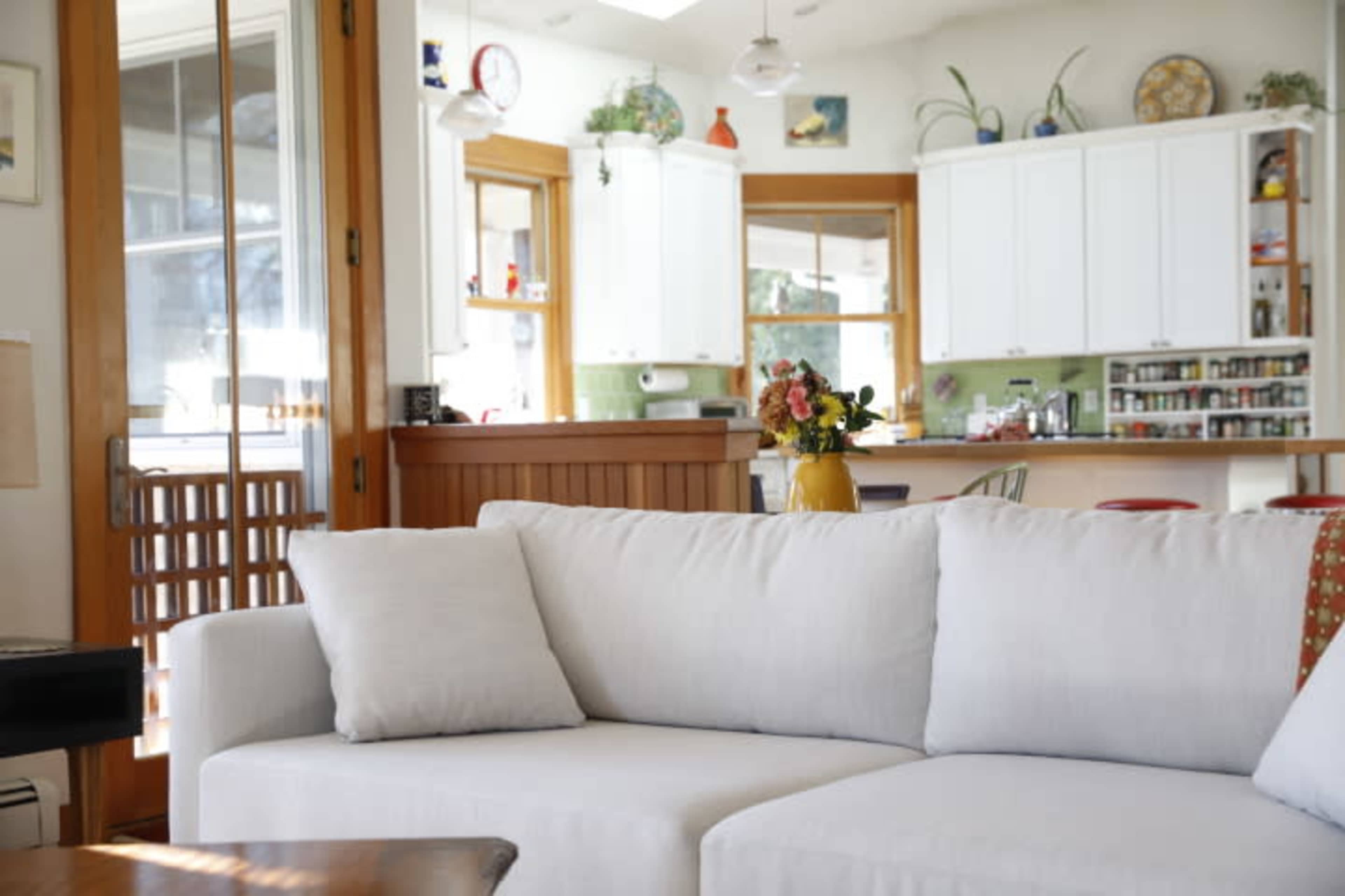 A light-colored sofa is positioned in a living room that opens to a kitchen with white cabinets and a variety of shelves and containers visible.