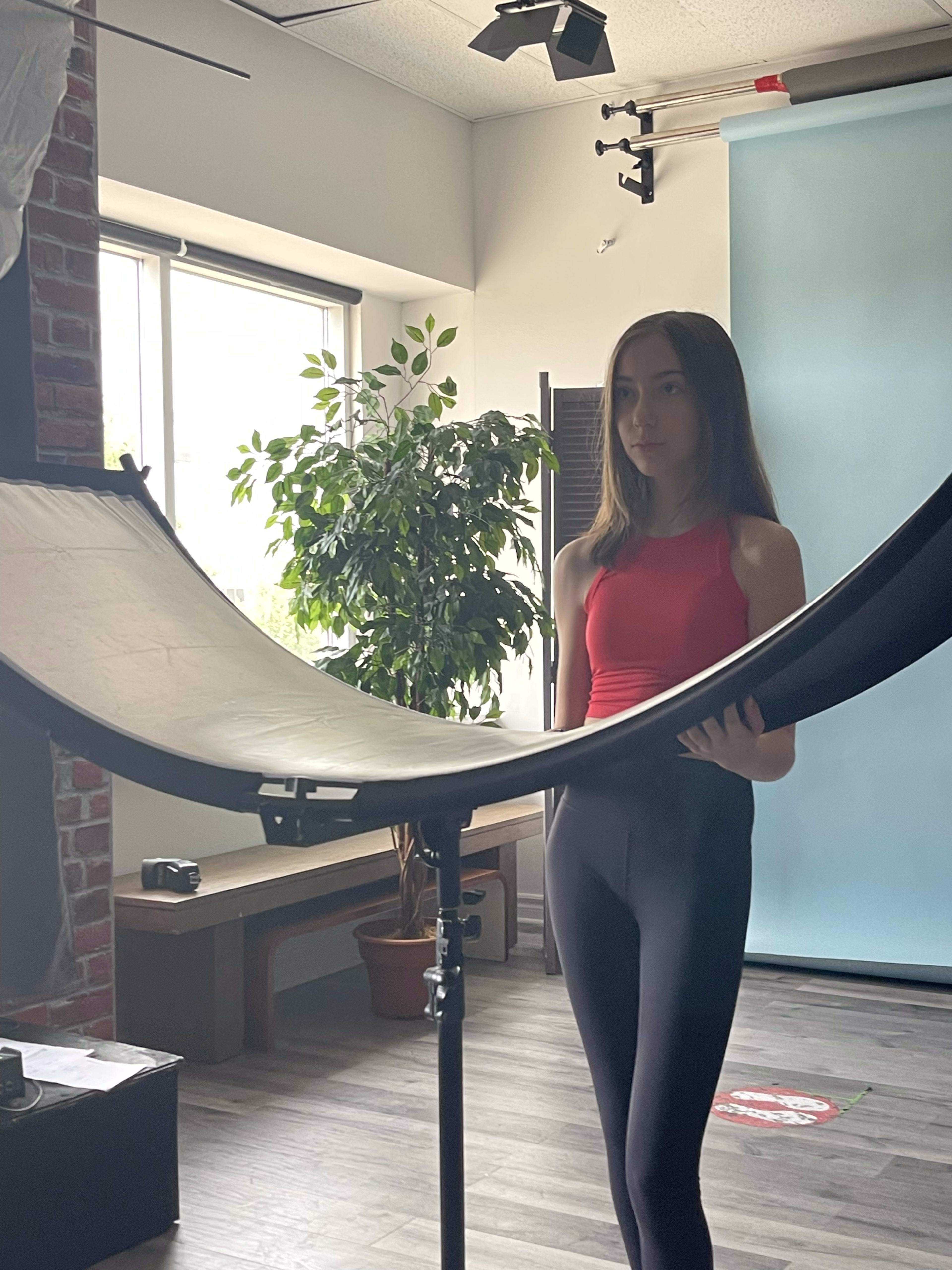 A young person in a red top stands beside a large reflector in a studio setting with a plant in the background.