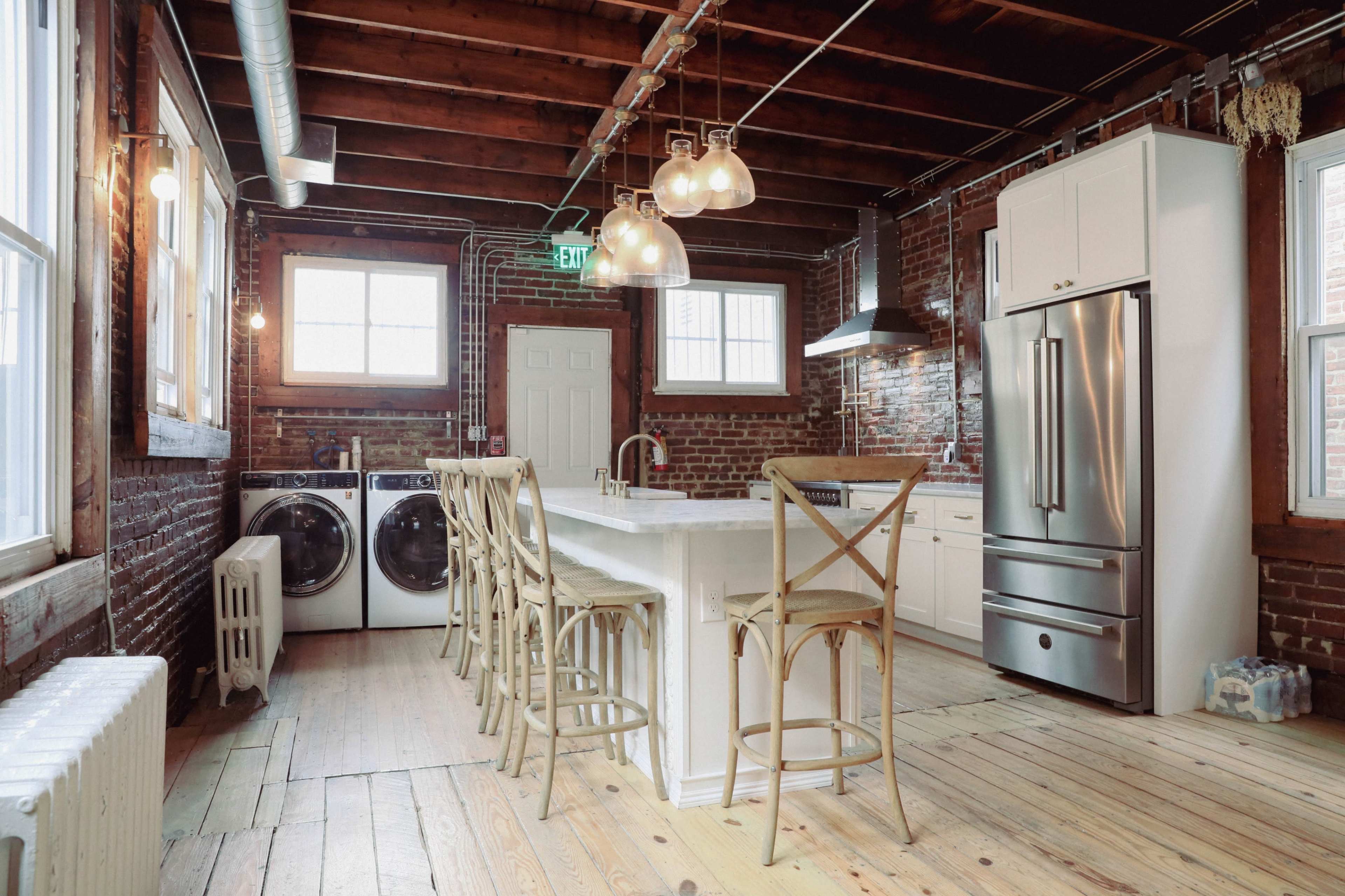 A modern kitchen with exposed brick walls, a large island with seating, and a washer and dryer located beside the refrigerator.