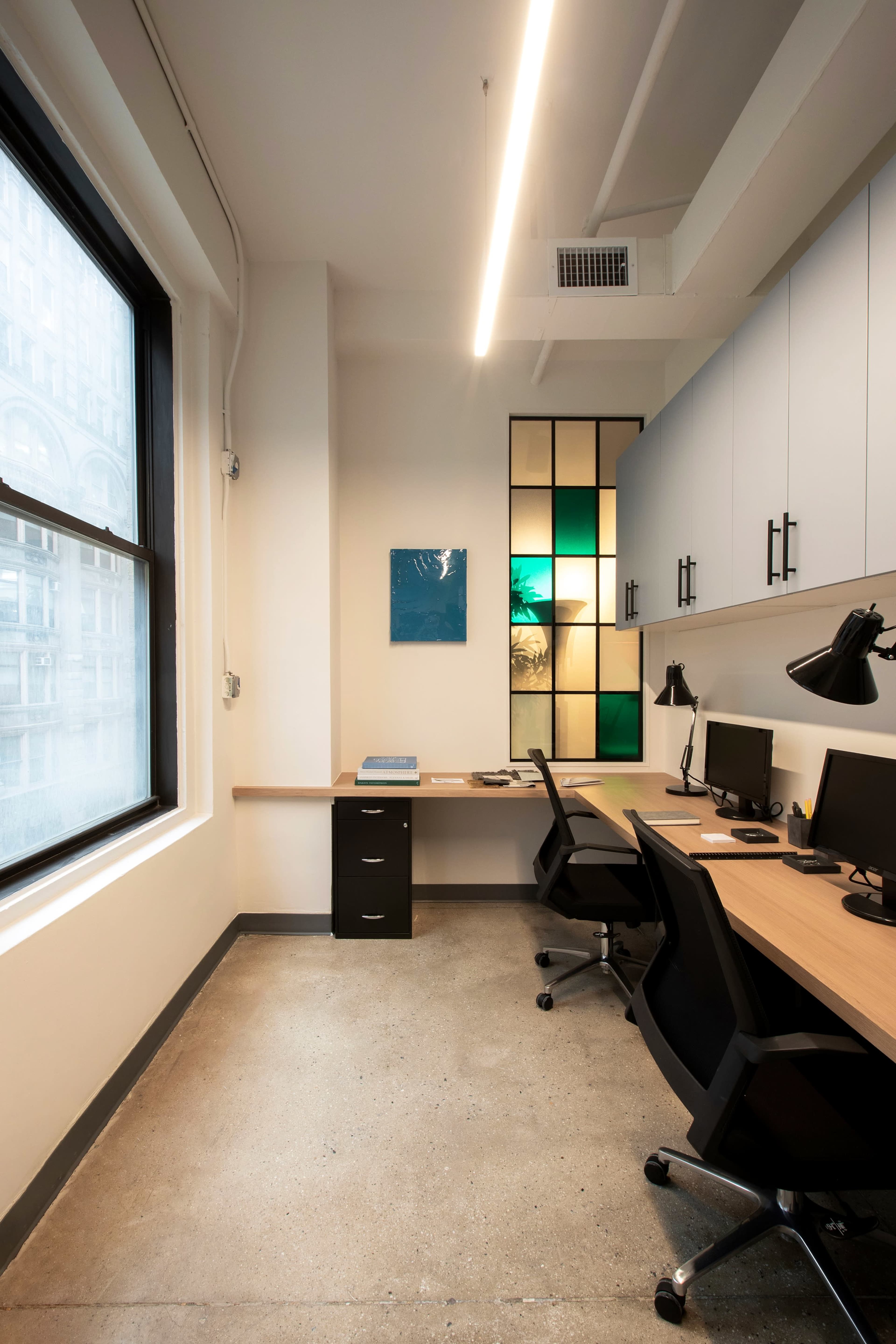 A modern office space with two desks, computers, and a large window letting in natural light.