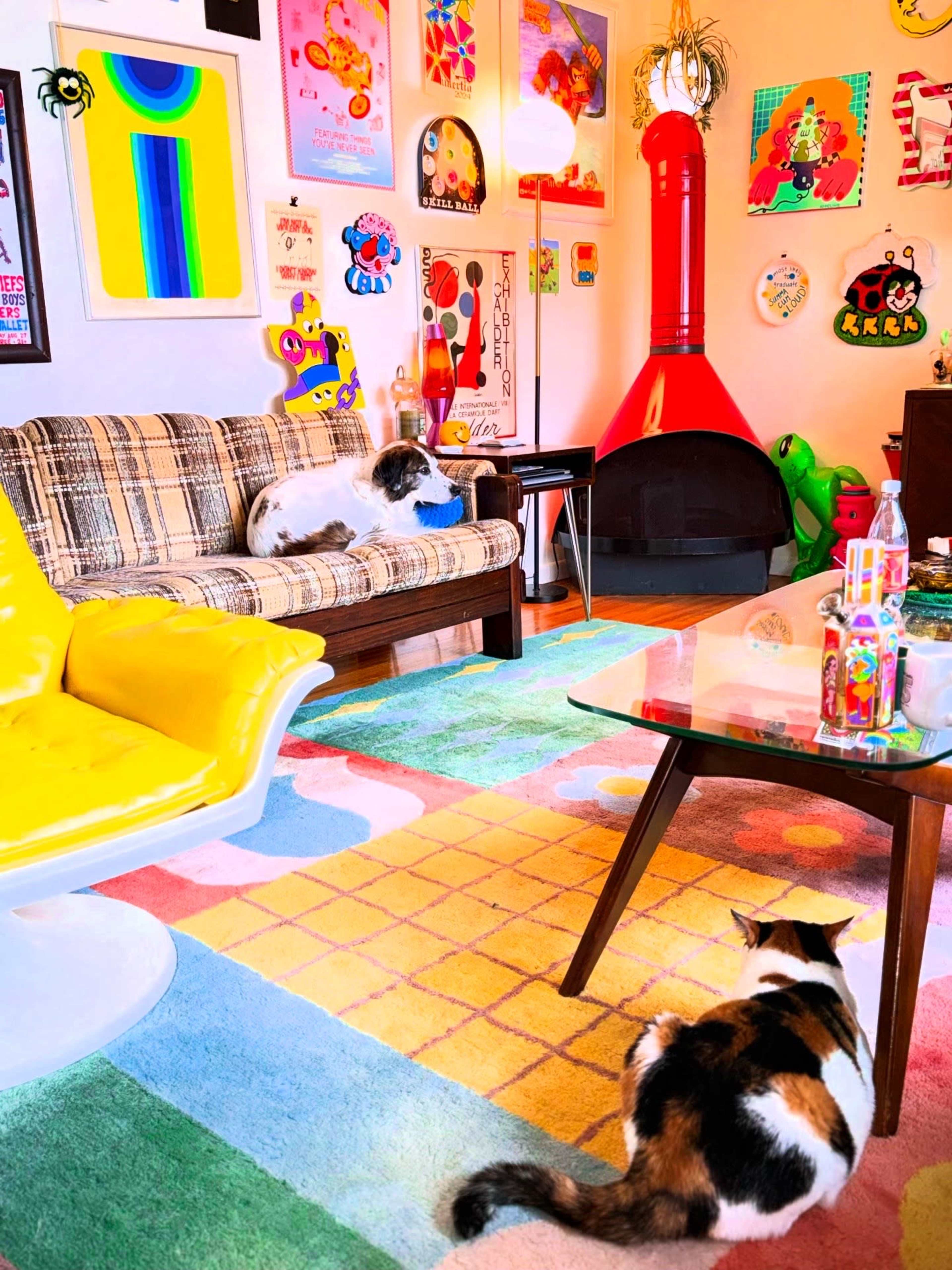 The room features a colorful, eclectic interior with a plaid sofa, a bright yellow chair, and two cats positioned on a patterned rug.