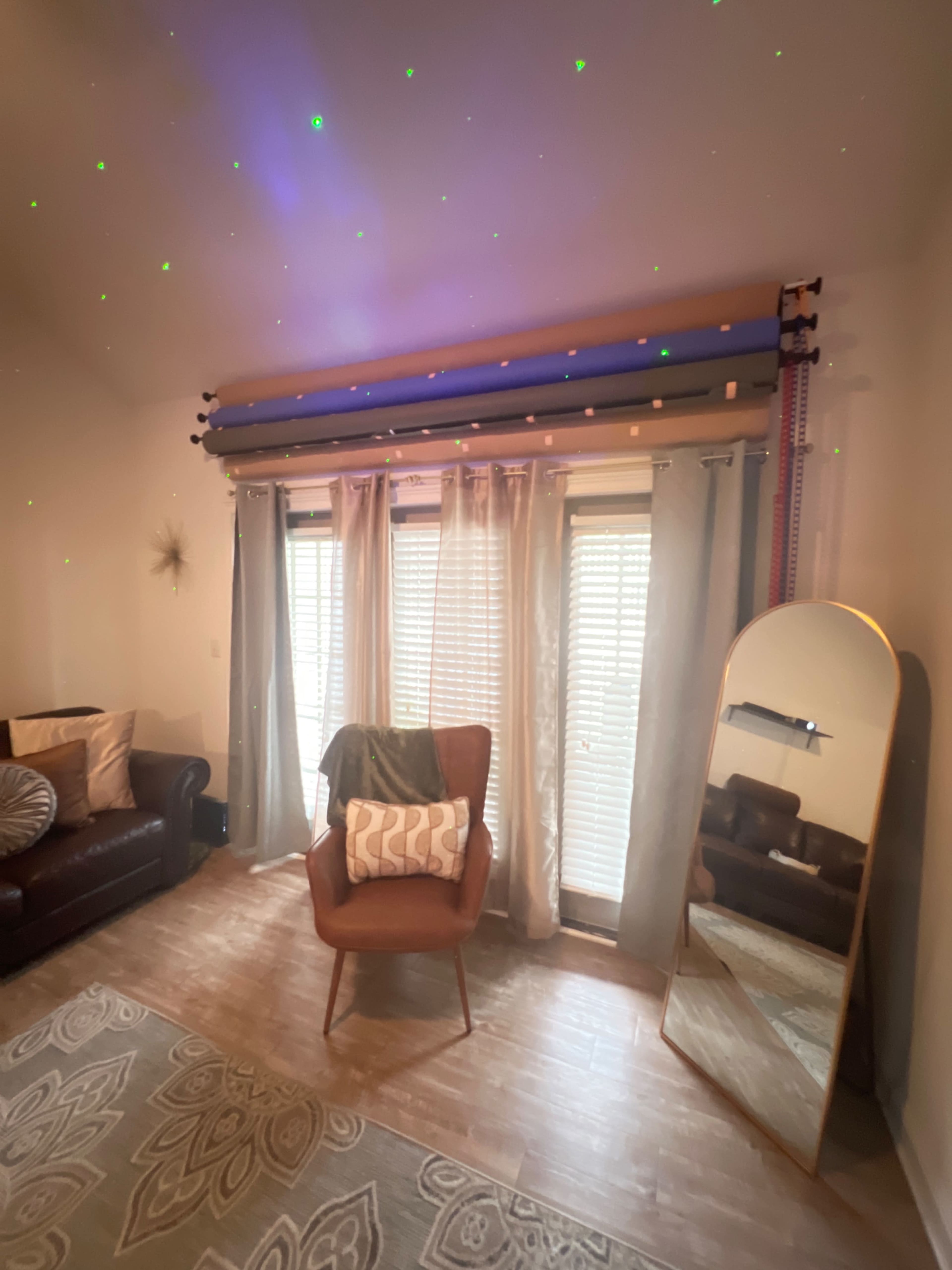 A cozy living room features a brown leather couch, a chair with a patterned cushion, sheer curtains, and a large mirror reflecting the space.