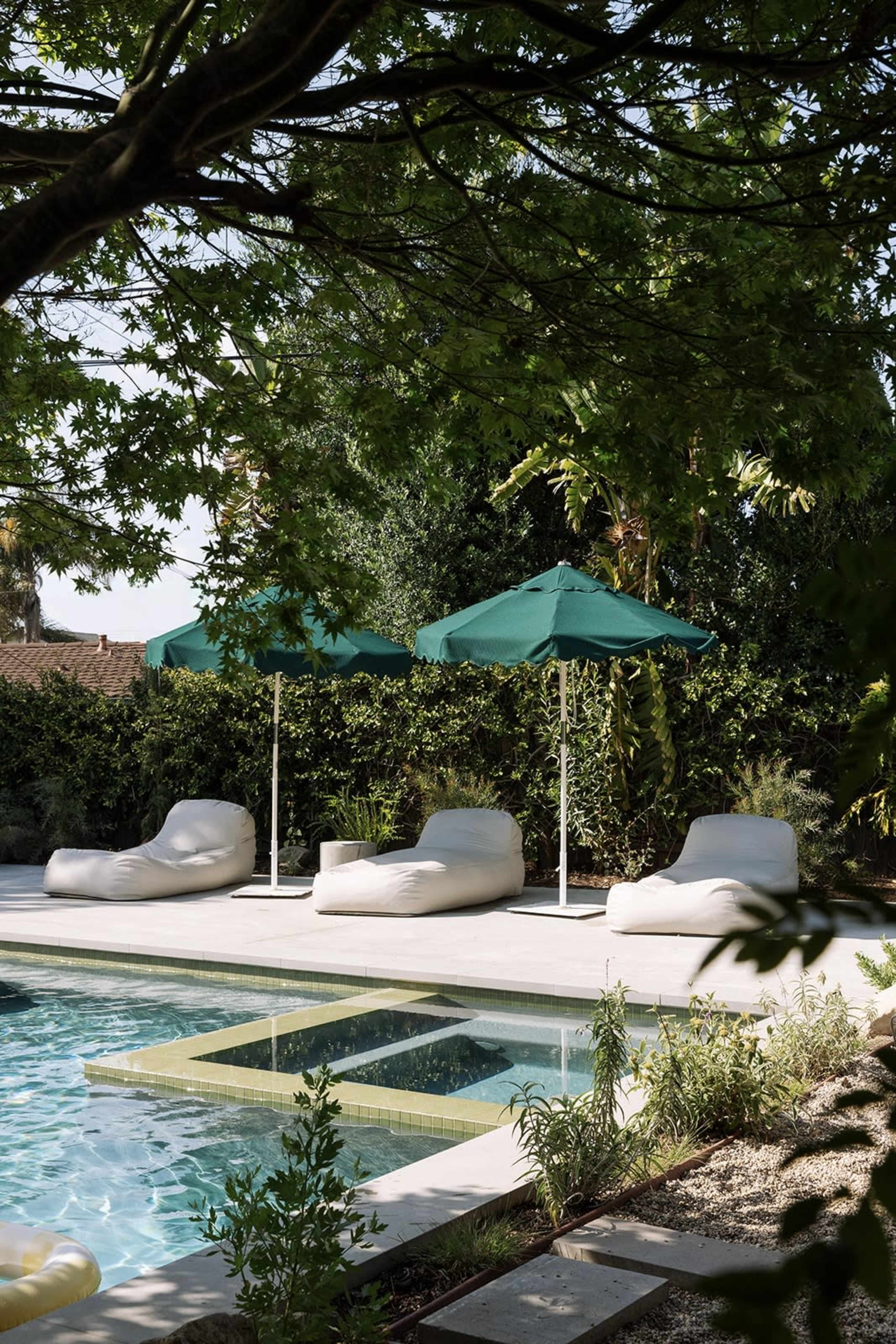 The image shows a pool area with three light-colored bean bag chairs and two green umbrellas surrounded by lush greenery.