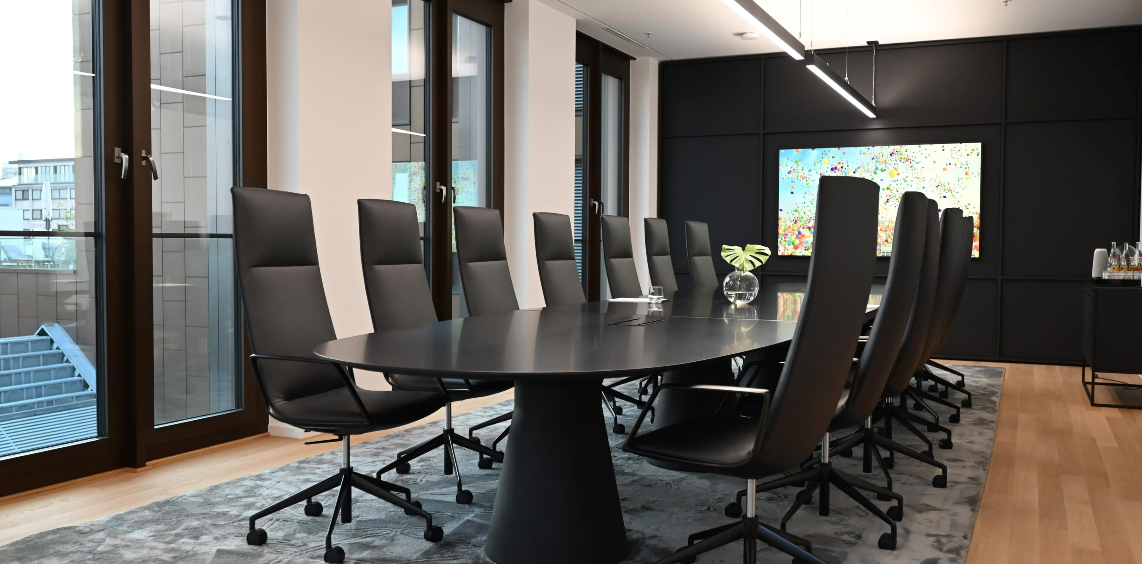 A modern conference room features a large oval table surrounded by ergonomic chairs, with floor-to-ceiling windows and contemporary artwork on the walls.